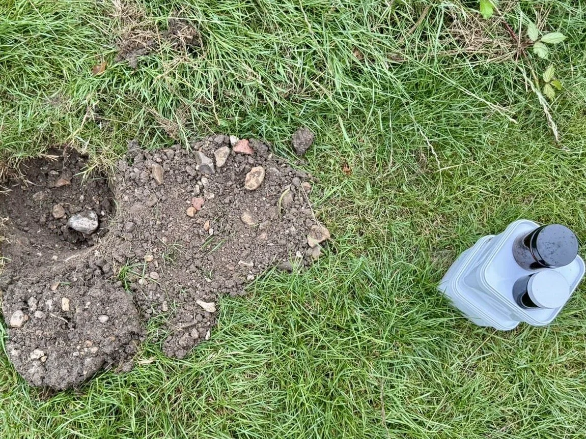 Freshly dug holes in the ground next to a container of small jars of ink or paint on grassy ground.