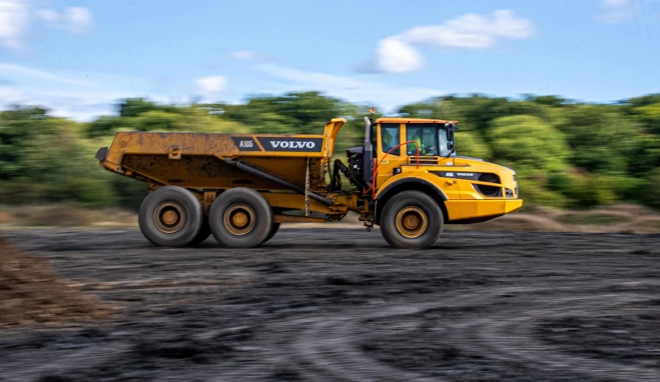 A yellow Volvo construction vehicle working on a large dirt or gravel surface, with green trees and a partly cloudy sky in the background.