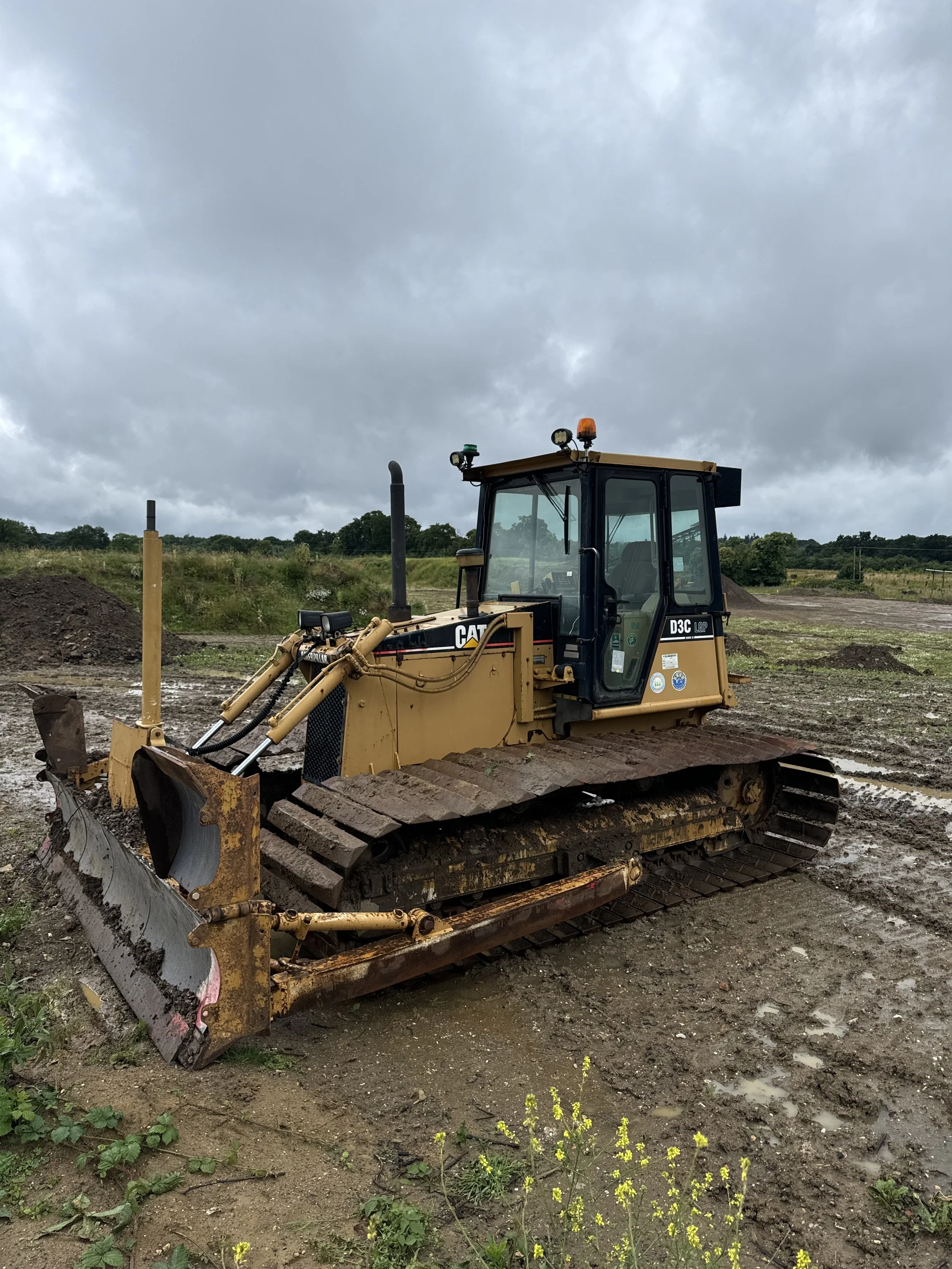 Yellow bulldozer on muddy terrain with cloudy sky and green landscape in the background.