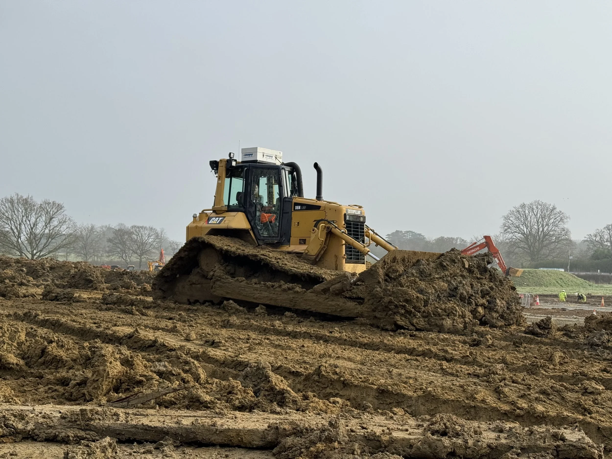 A yellow bulldozer working on a construction site, pushing dirt, with other construction equipment and workers in the background.
