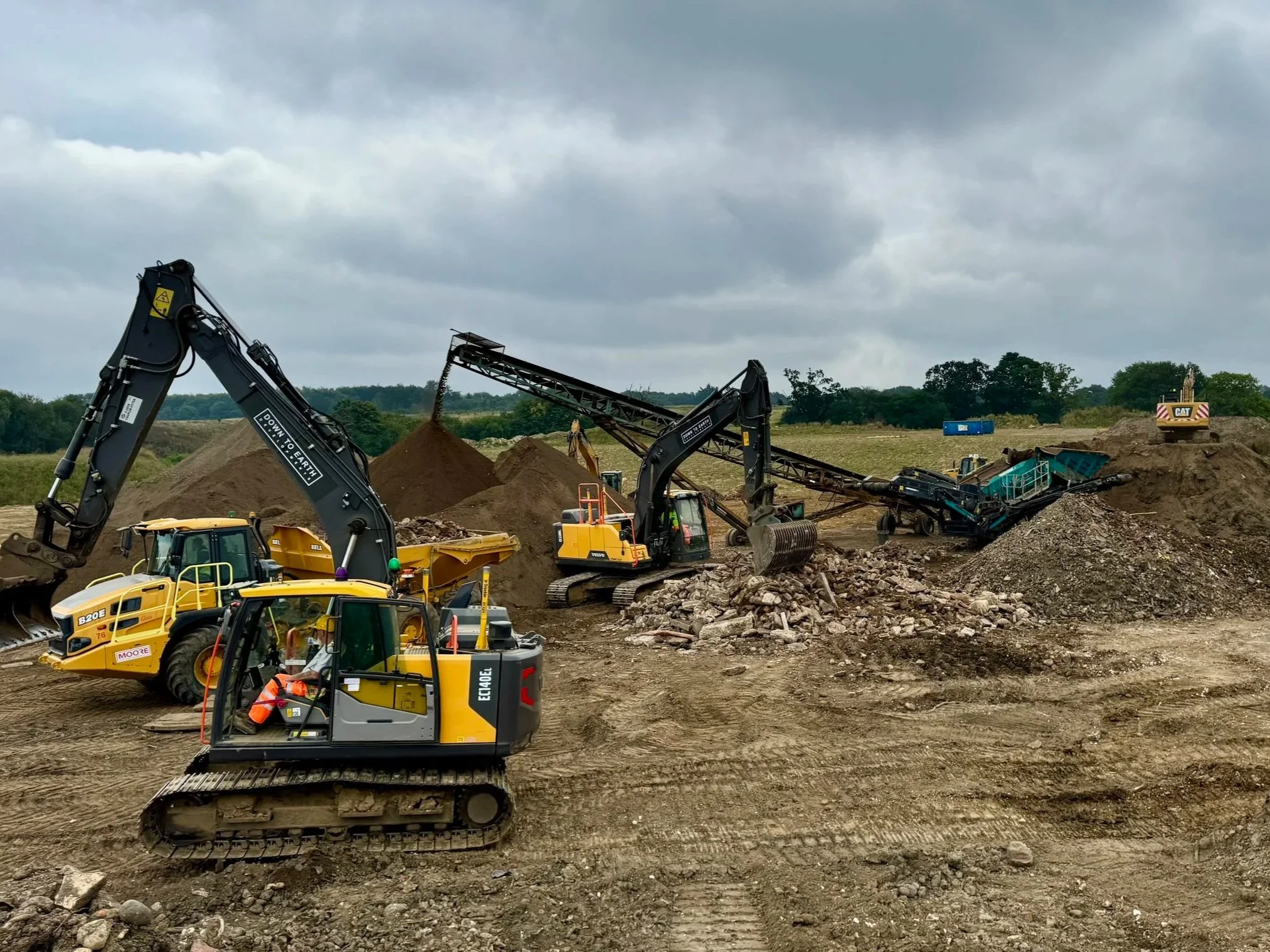 Construction site with multiple excavators moving dirt and rocks under a cloudy sky.