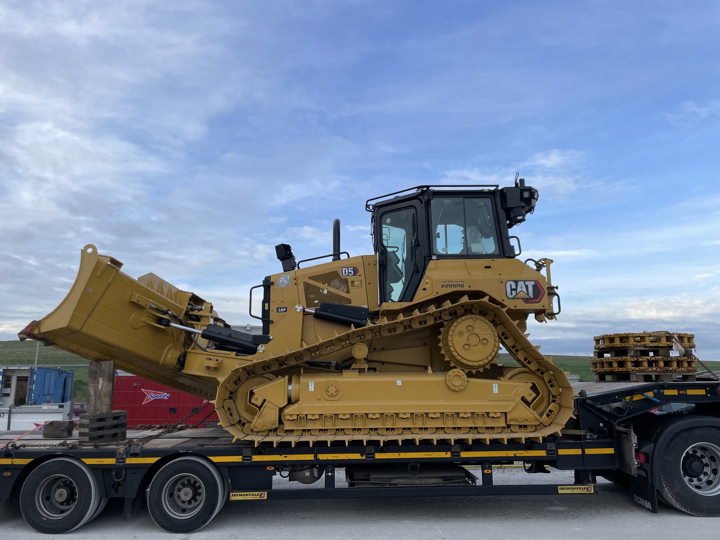 A yellow Caterpillar bulldozer loaded on a flatbed truck with a background of blue sky and some clouds.