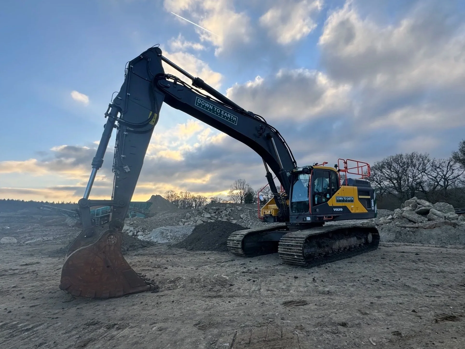 A black and yellow excavator at a construction site with rocks and dirt, under a partly cloudy sky during sunset.