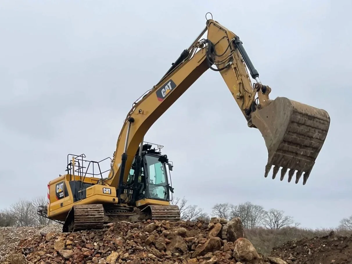 A yellow Caterpillar excavator with a large bucket attachment is on a pile of dirt and rocks, working on a construction site under an overcast sky.