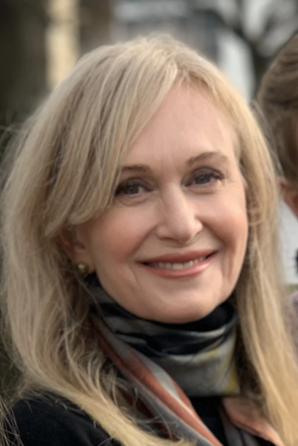 A smiling blonde woman with blue eyes, wearing a black top and a patterned scarf.