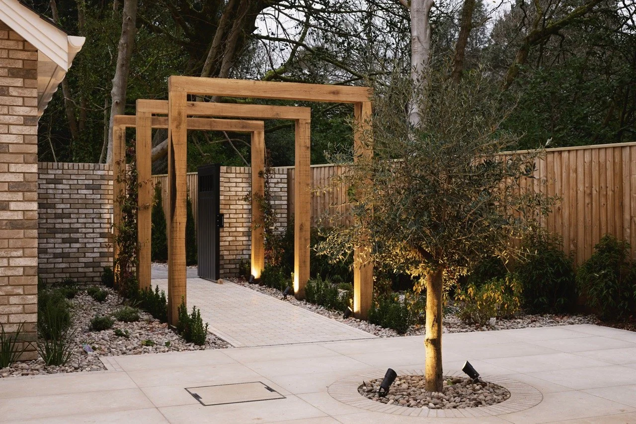 A landscaped backyard with a paved walkway, a small tree with uplighting, a wooden pergola frame, brick and wooden fencing, and garden lights.