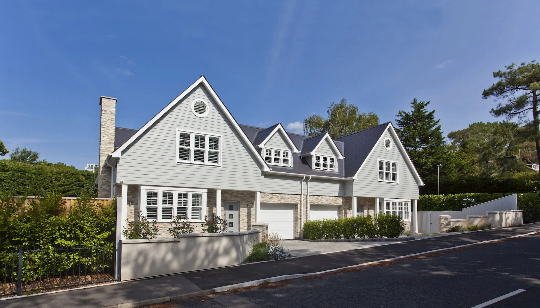 A modern, multi-story house situated in Poole with grey siding, white trim, and black roof shingles, featuring several gabled roofs, large windows with shutters, a driveway with two garages, and landscapedfront yard, set against a backdrop of trees.
