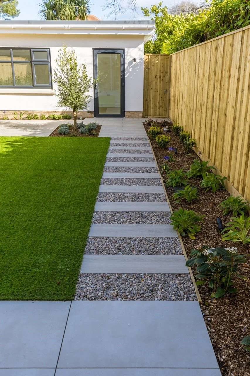 A modern backyard with a concrete walkway, gravel strips, a grassy lawn, a small tree, flower beds on the right, and a wooden fence.