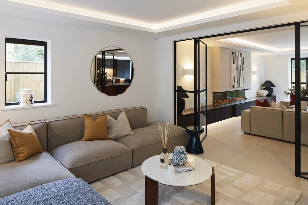 Modern living room with beige sofas, decorative pillows, a round white coffee table, and a wall-mounted fireplace, separated by glass sliding doors.