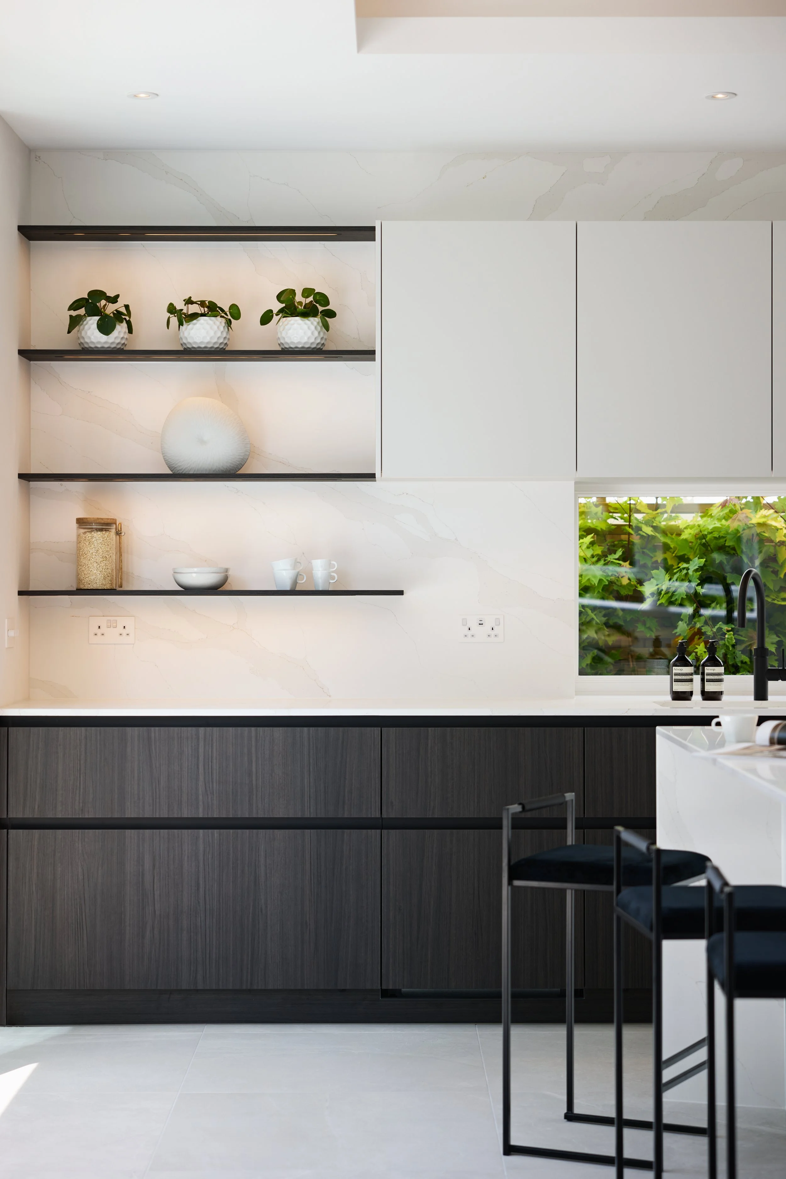 Modern kitchen with dark lower cabinets, white upper cabinets, white marble backsplash, open black shelves with plants and decor, window revealing greenery outside, black bar stools at white island, bottles and cups on counter.