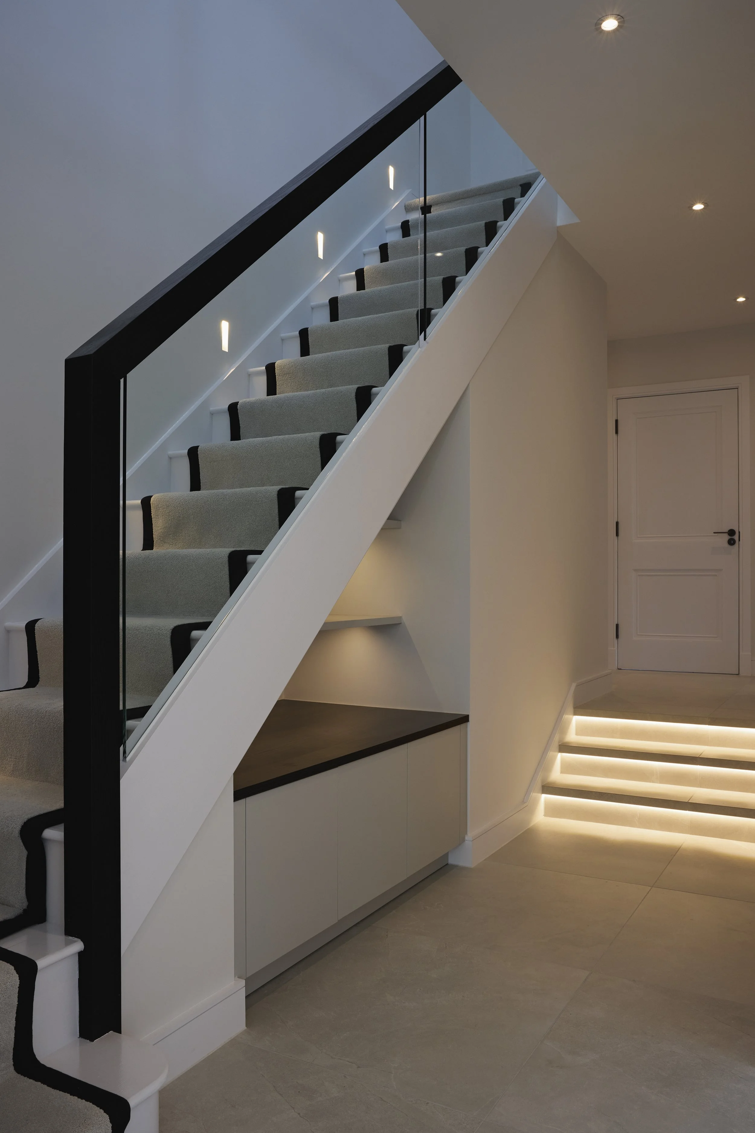 Modern staircase with beige carpet, black trim, glass railing, and built-in lighting in a contemporary home interior.