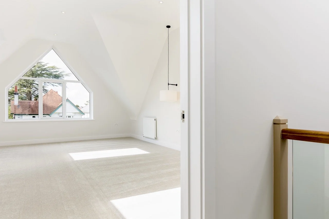Empty attic room with a large triangular window, angled ceiling, hanging light fixture, white walls, and beige carpet.