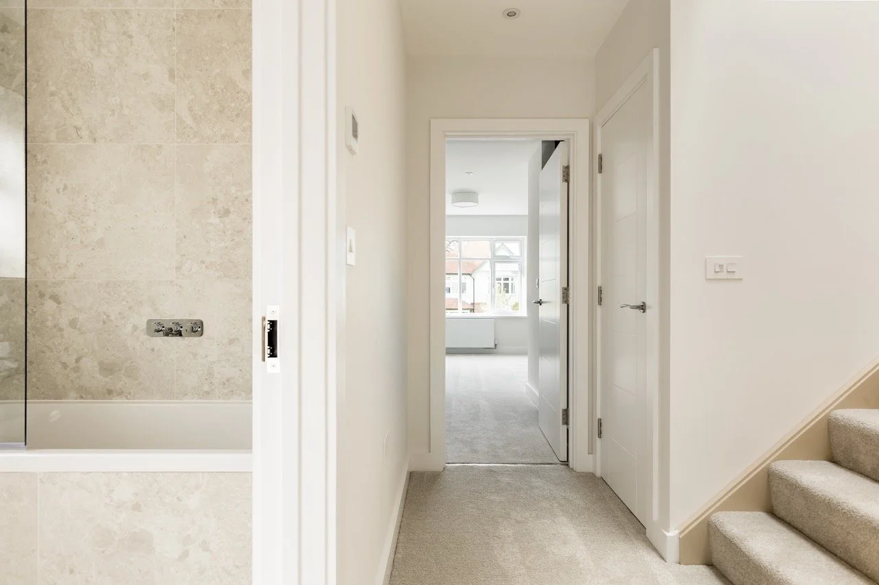 Interior view of a hallway leading to a bright room with window, partially visible bathroom with tiled shower on the left, and staircase on the right.