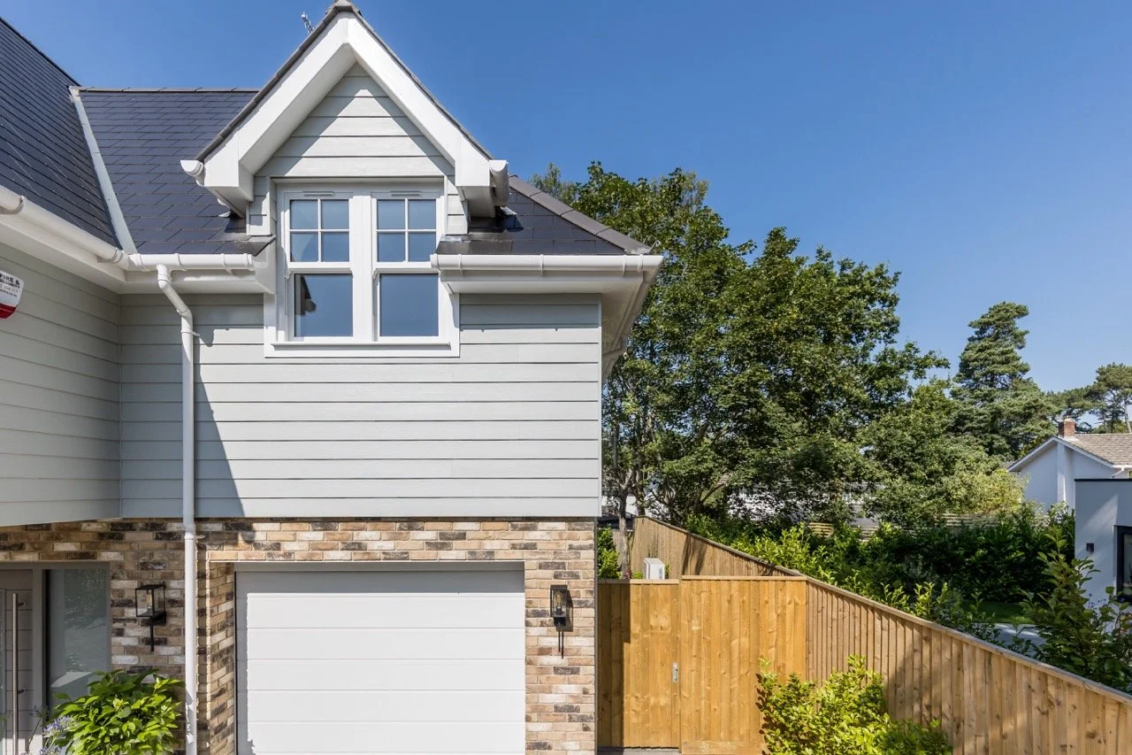 Modern two-story house with grey siding, brick accents, a white garage door, and a gabled window on the upper level. There is a wooden fence on the right and green trees in the background under a clear blue sky.