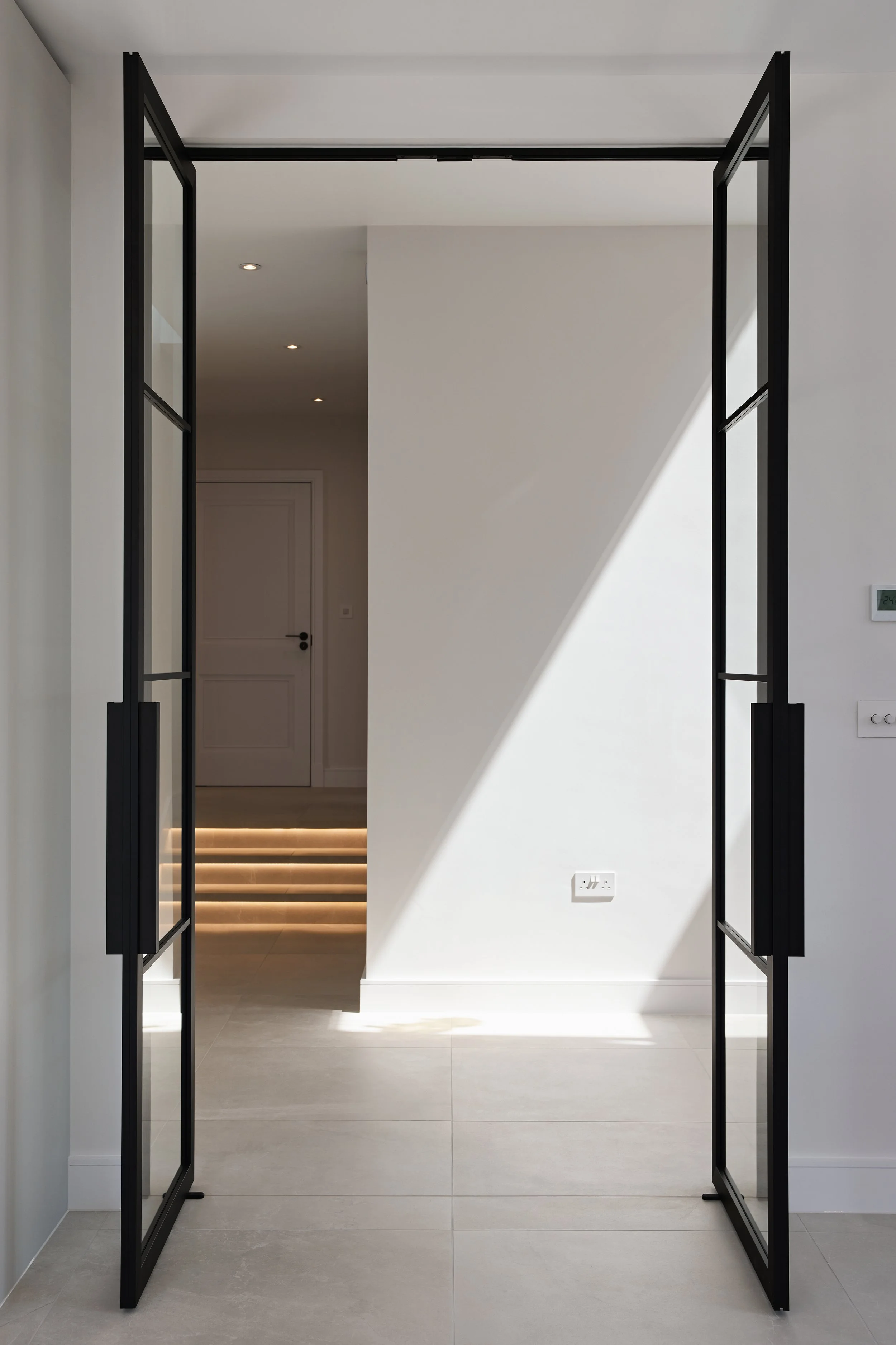 Modern doorway with black-framed glass doors leading into a bright interior with white walls, tiled floor, and illuminated steps on a staircase in the background.