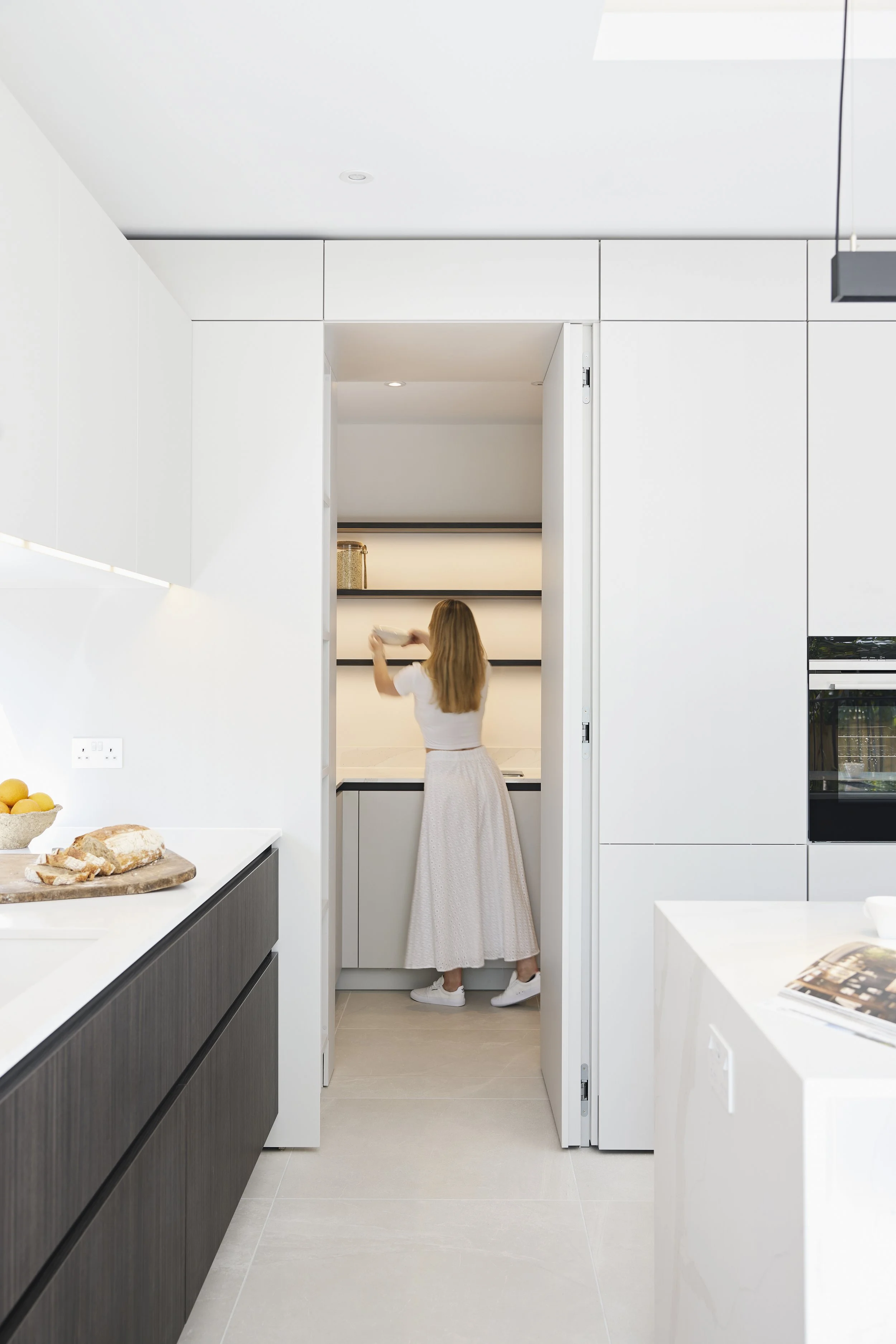 A woman with long hair wearing a white t-shirt and a long skirt reaches for a shelf in a minimalist white kitchen.
