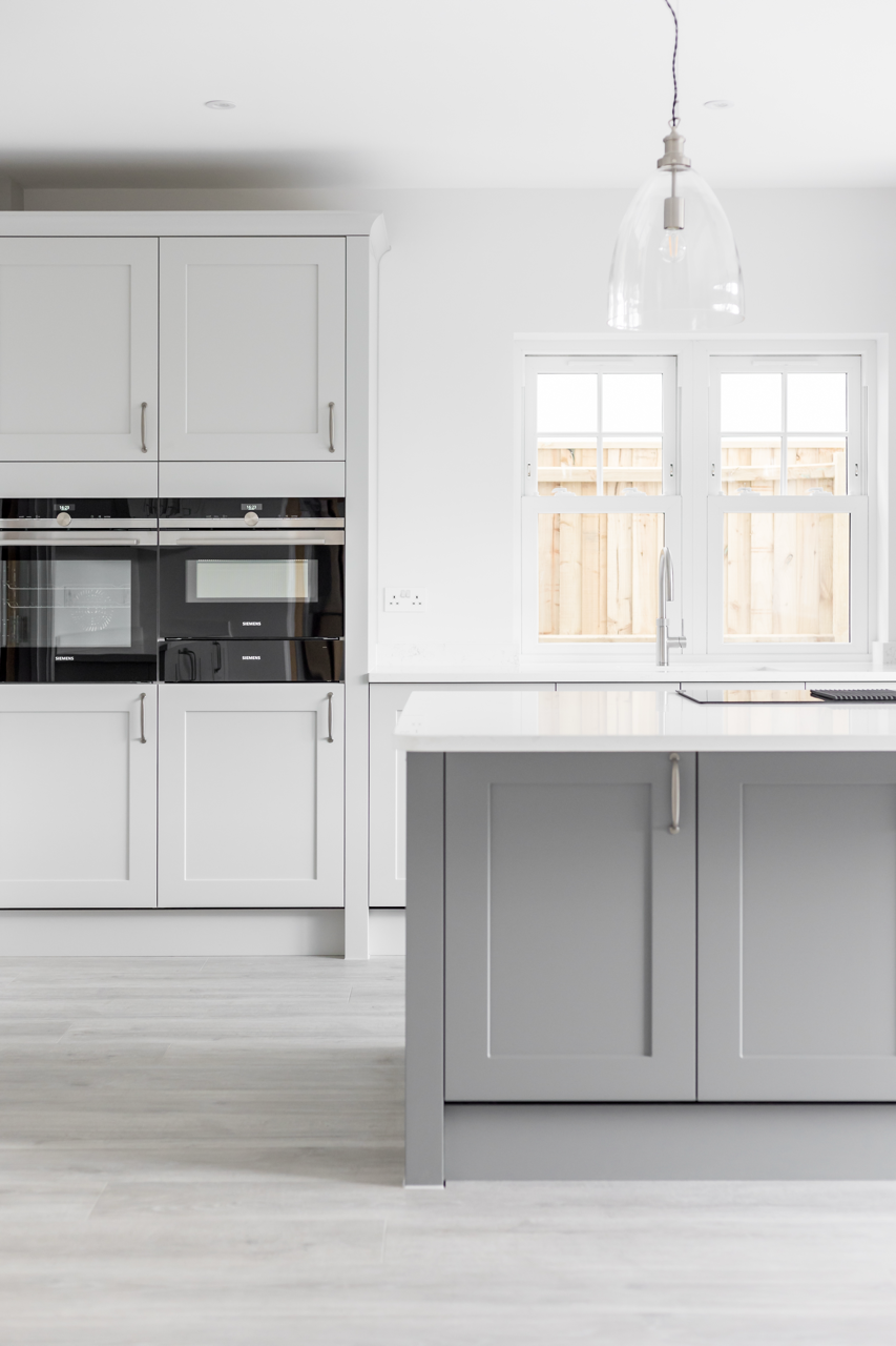Modern minimalist kitchen with white cabinets, a gray kitchen island, and double windows overlooking a wooden fence.