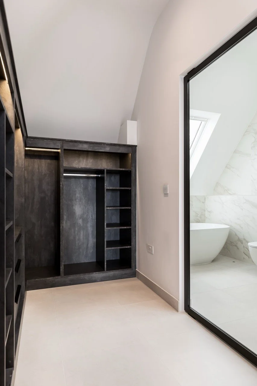 Empty walk-in closet with black shelving and hanging rod, adjacent to a modern bathroom with a freestanding bathtub, marble walls, and a skylight