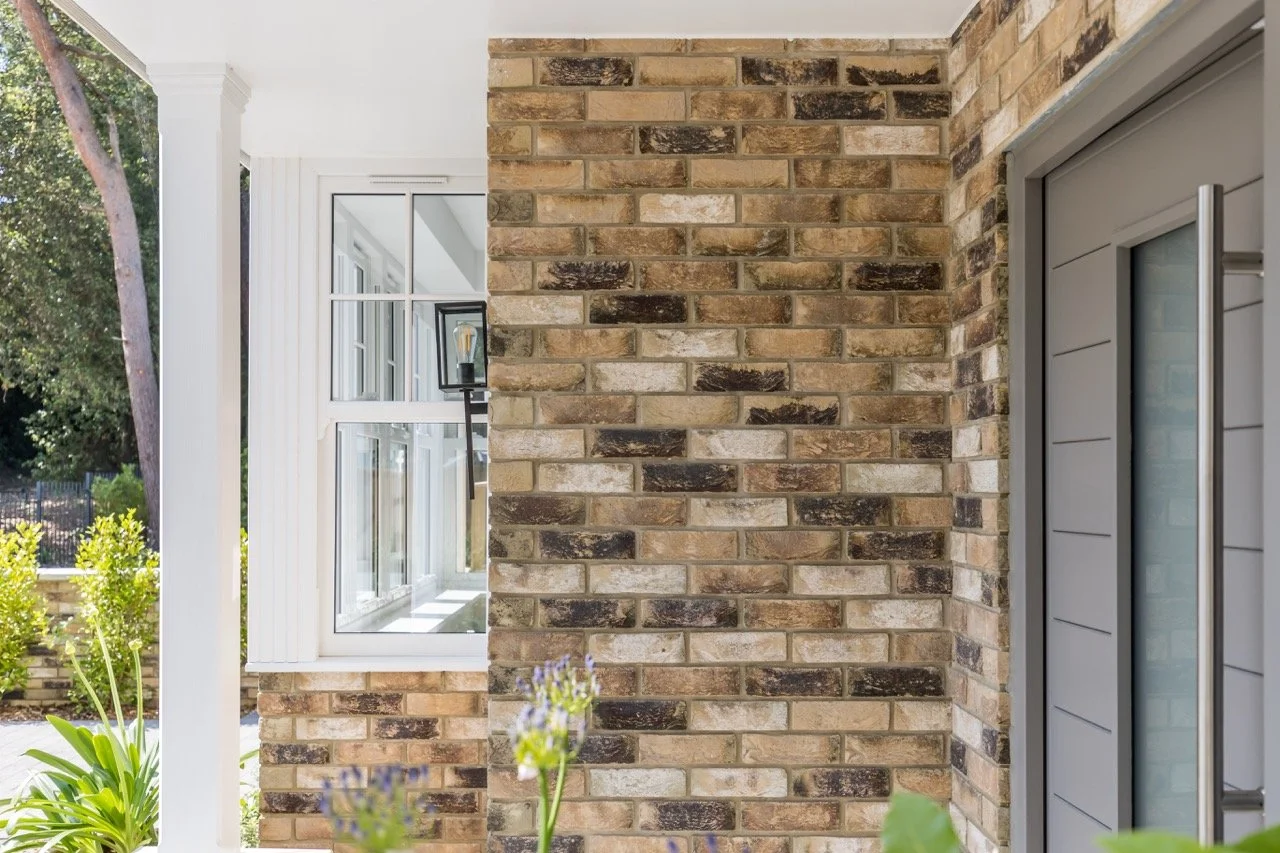 Close-up of a brick wall on a house exterior, with a window and a sliding door, and some greenery in the foreground.