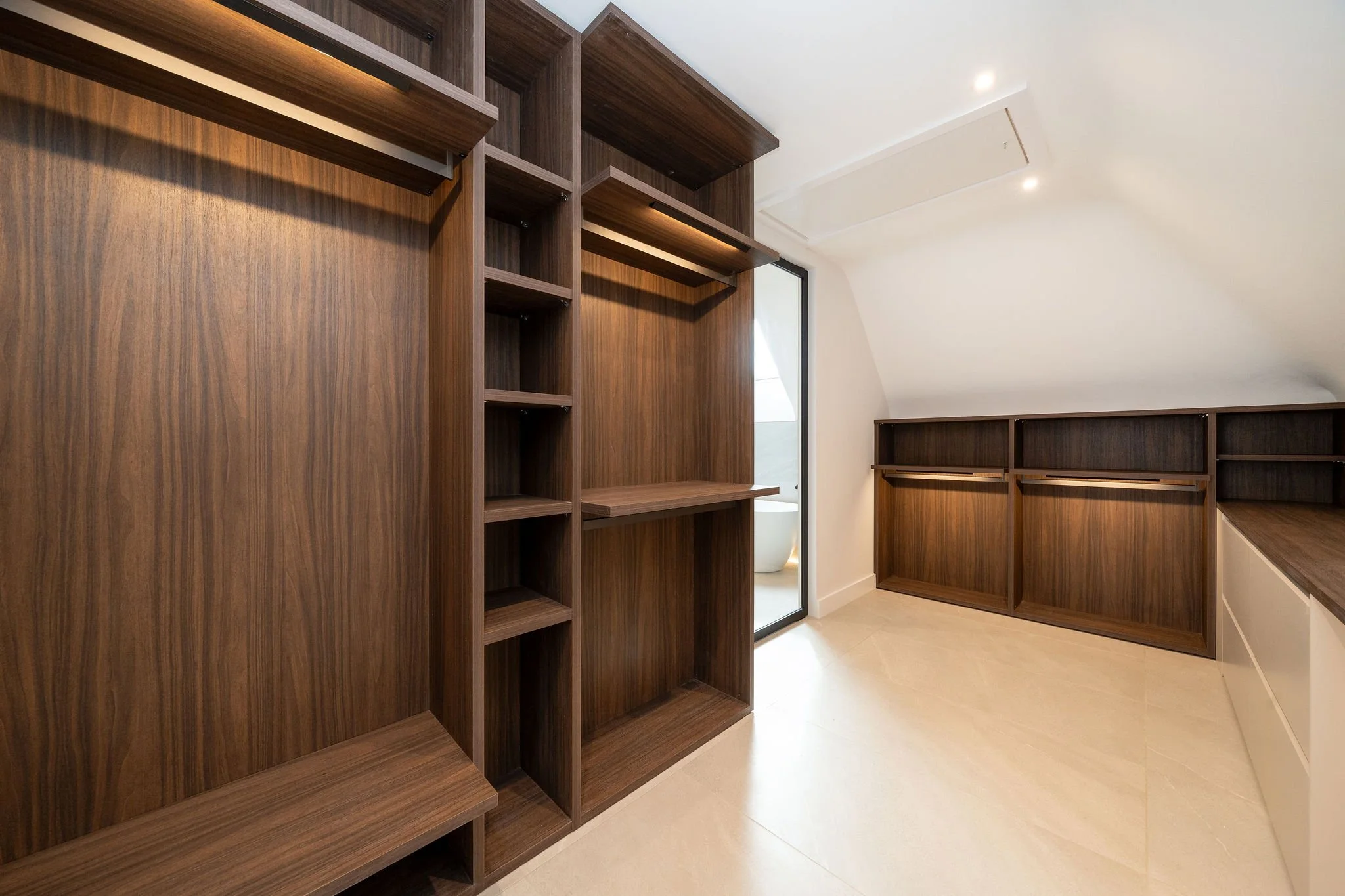 Empty walk-in closet with dark wood shelves and hooks, light-colored tiled floor, and a glass door leading to a bathroom with a bathtub.