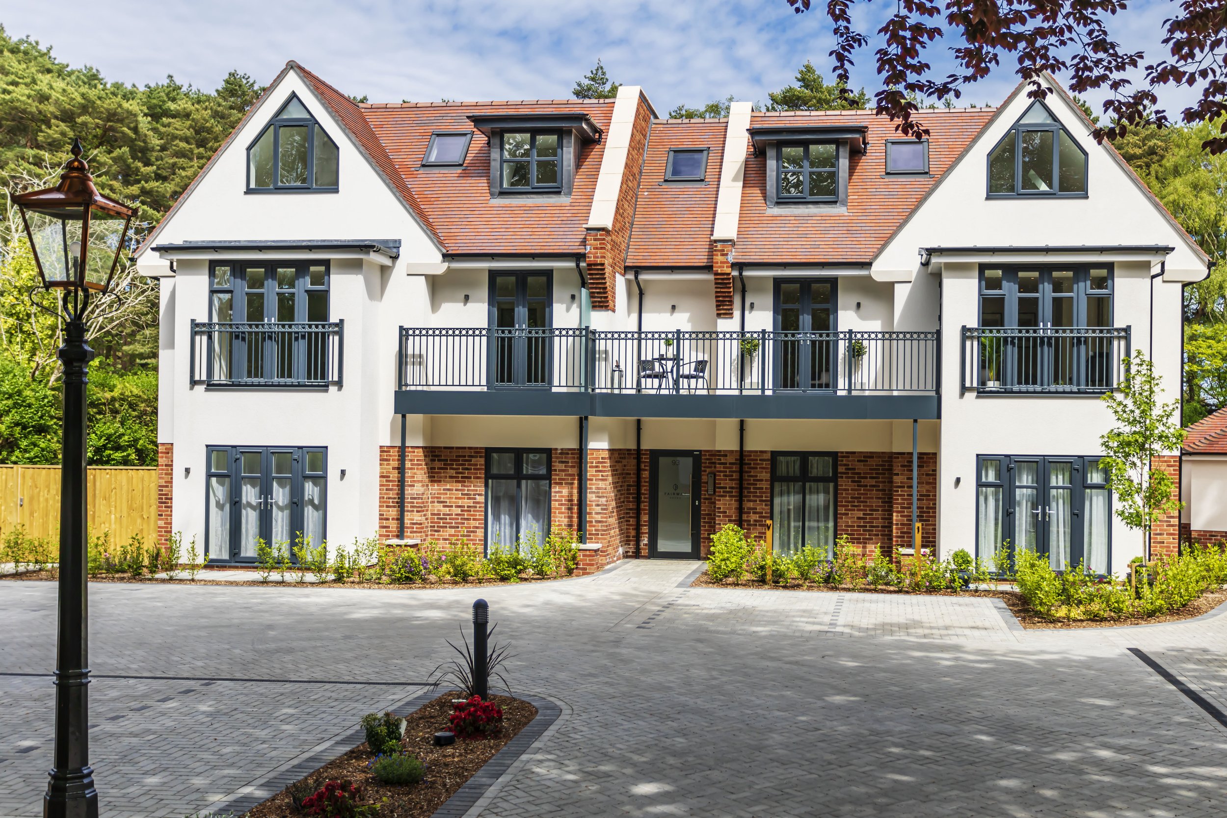 Modern multi-story house with white exterior, red-tile roof, and multiple windows, surrounded by trees and a paved driveway with landscaped flower beds.