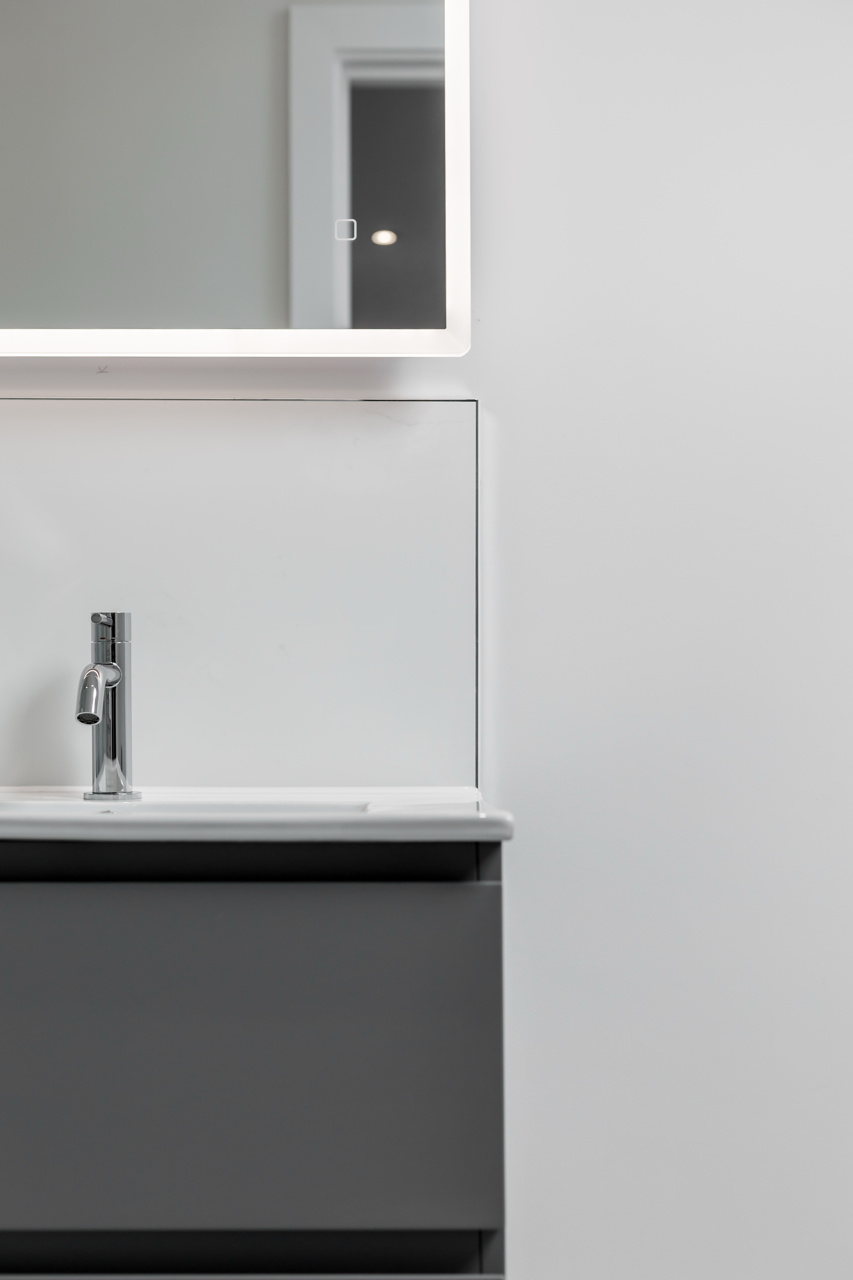 Close-up of a modern bathroom sink with chrome faucet, gray cabinet, white wall, and a mirror above the sink reflecting a doorway and light fixture.