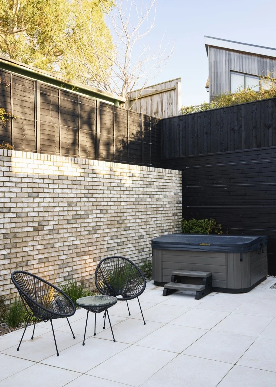 Outdoor patio area featuring two black wire chairs with a small matching side table, a hot tub with a cover, white tiled flooring, and a backdrop of a white brick wall and black wooden fencing.
