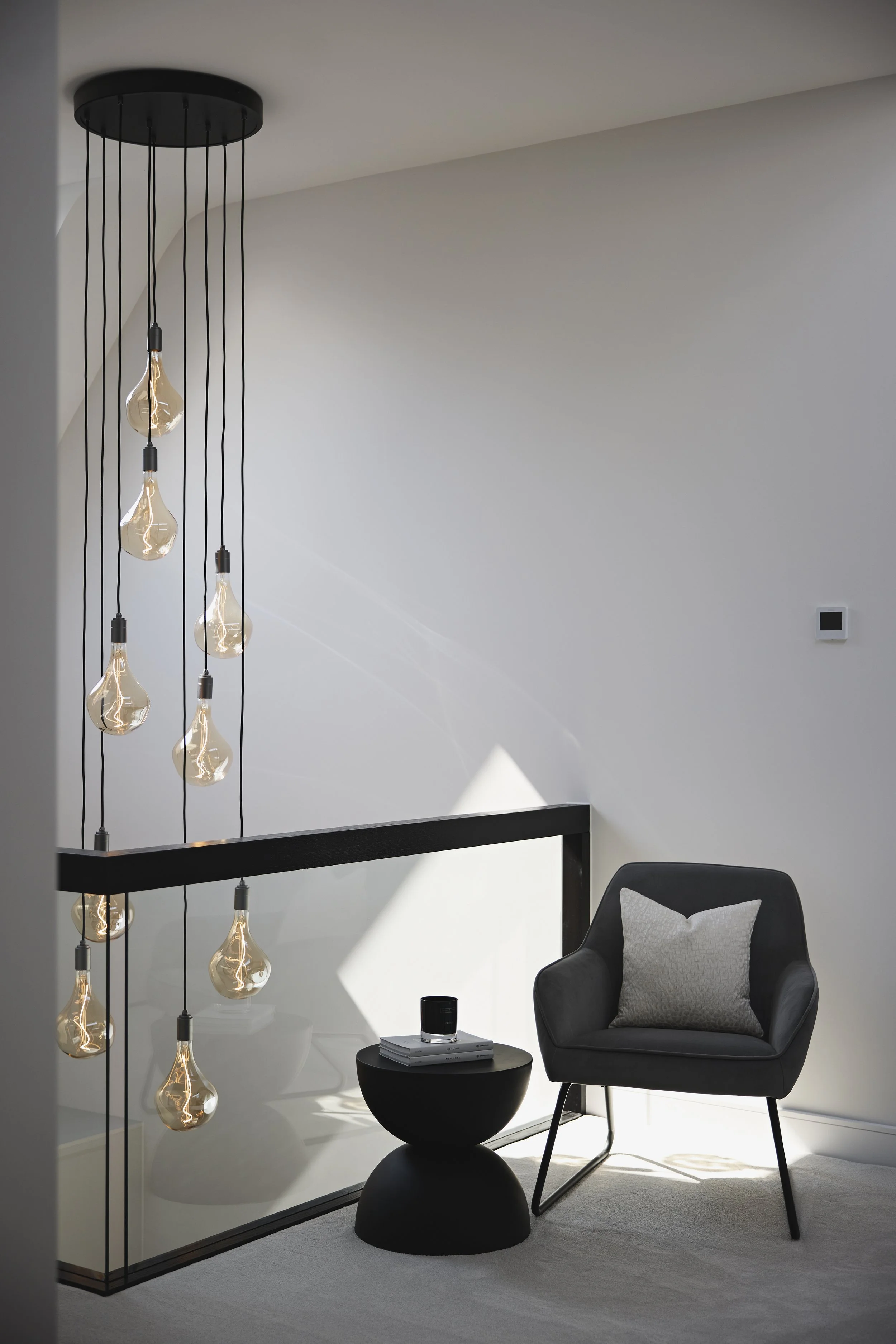 Modern interior space with hanging pendant lights, a black armchair with a cushion, a round black side table with books and a speaker, and a white wall with a thermostat.