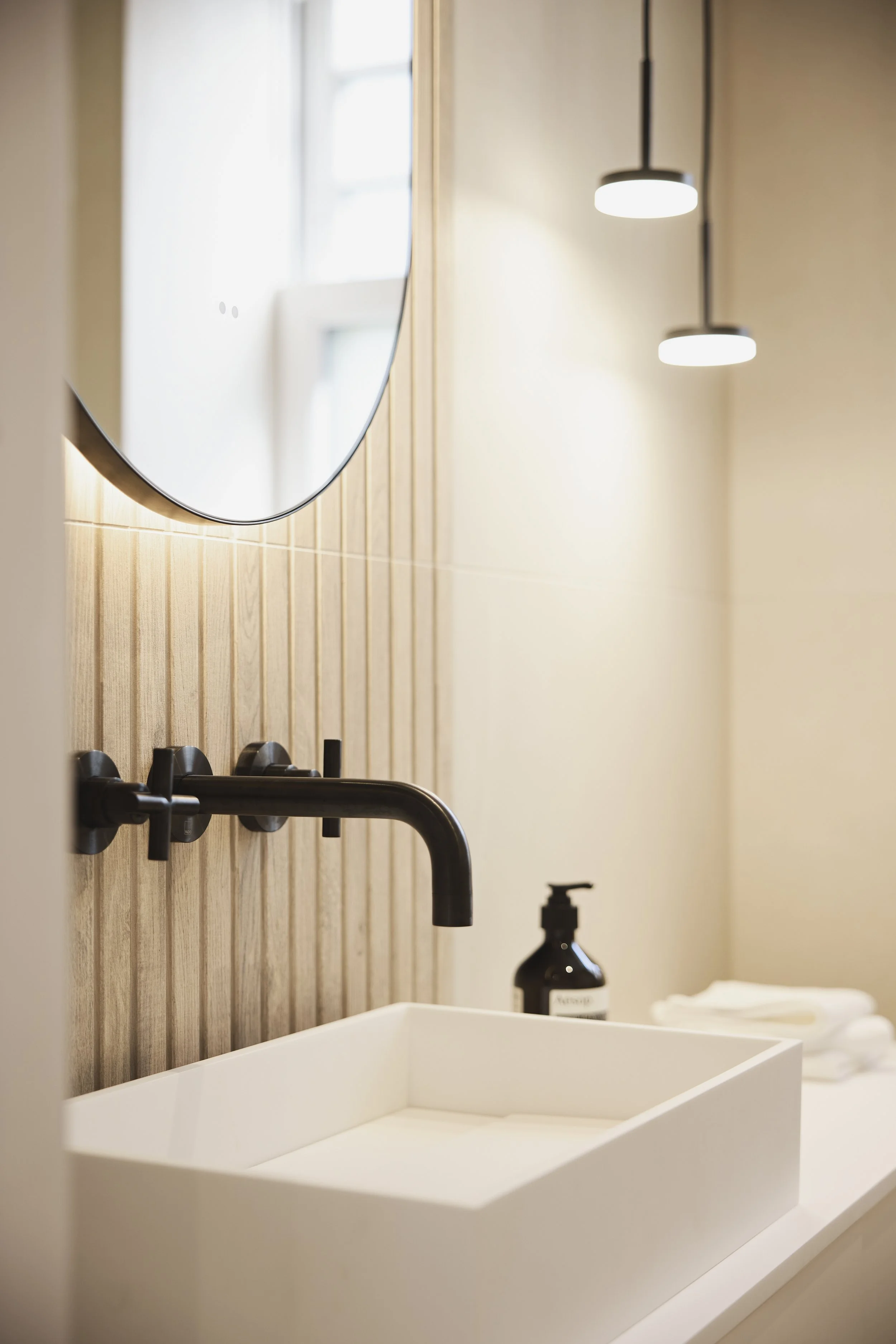 Modern bathroom with a white rectangular sink, black wall-mounted faucet, round mirror, natural wood wall paneling, and hanging light fixtures