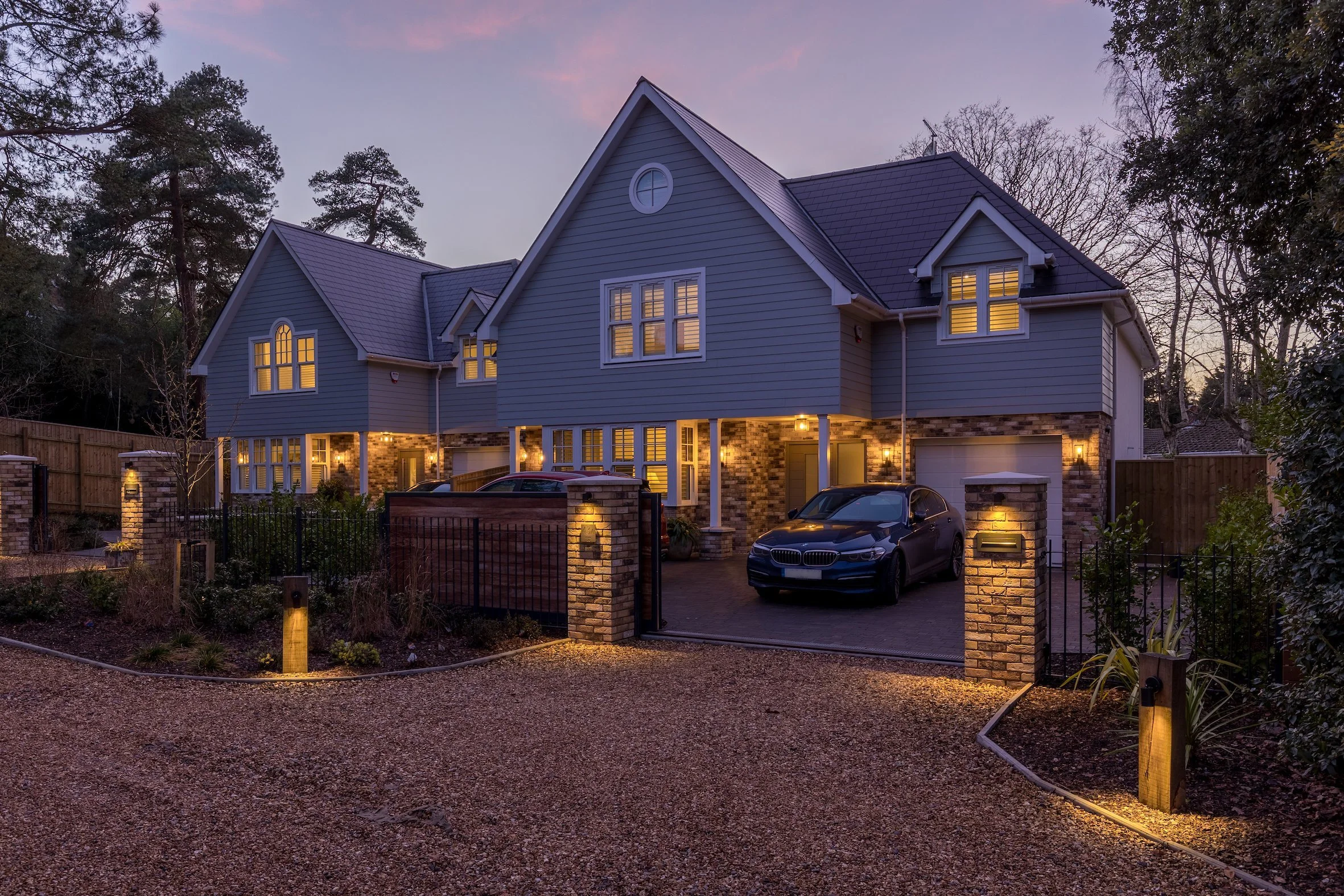 A new CGI render of two modern three-story houses located in the South Coast in Poole, Dorset with large glass windows and balconies, illuminated from within, surrounded by trees and greenery at dusk, with a sports car parked in front.