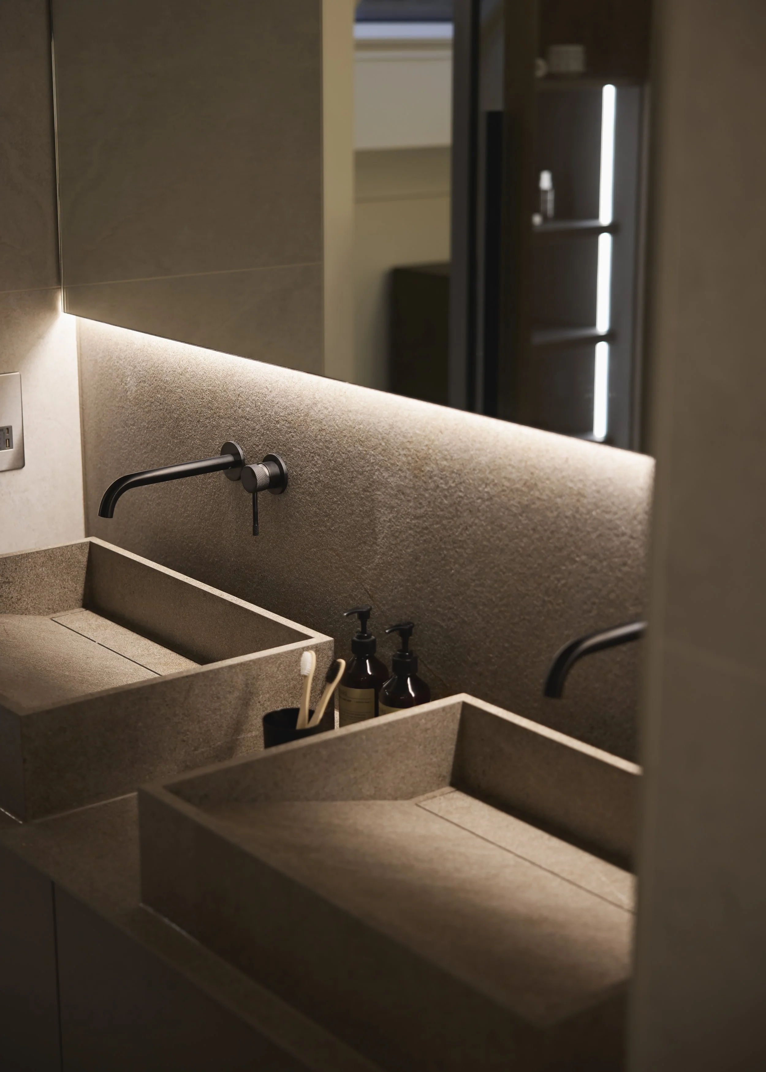 A modern bathroom double sink with black faucets, soap dispensers, toothbrushes, and a large mirror with soft lighting.