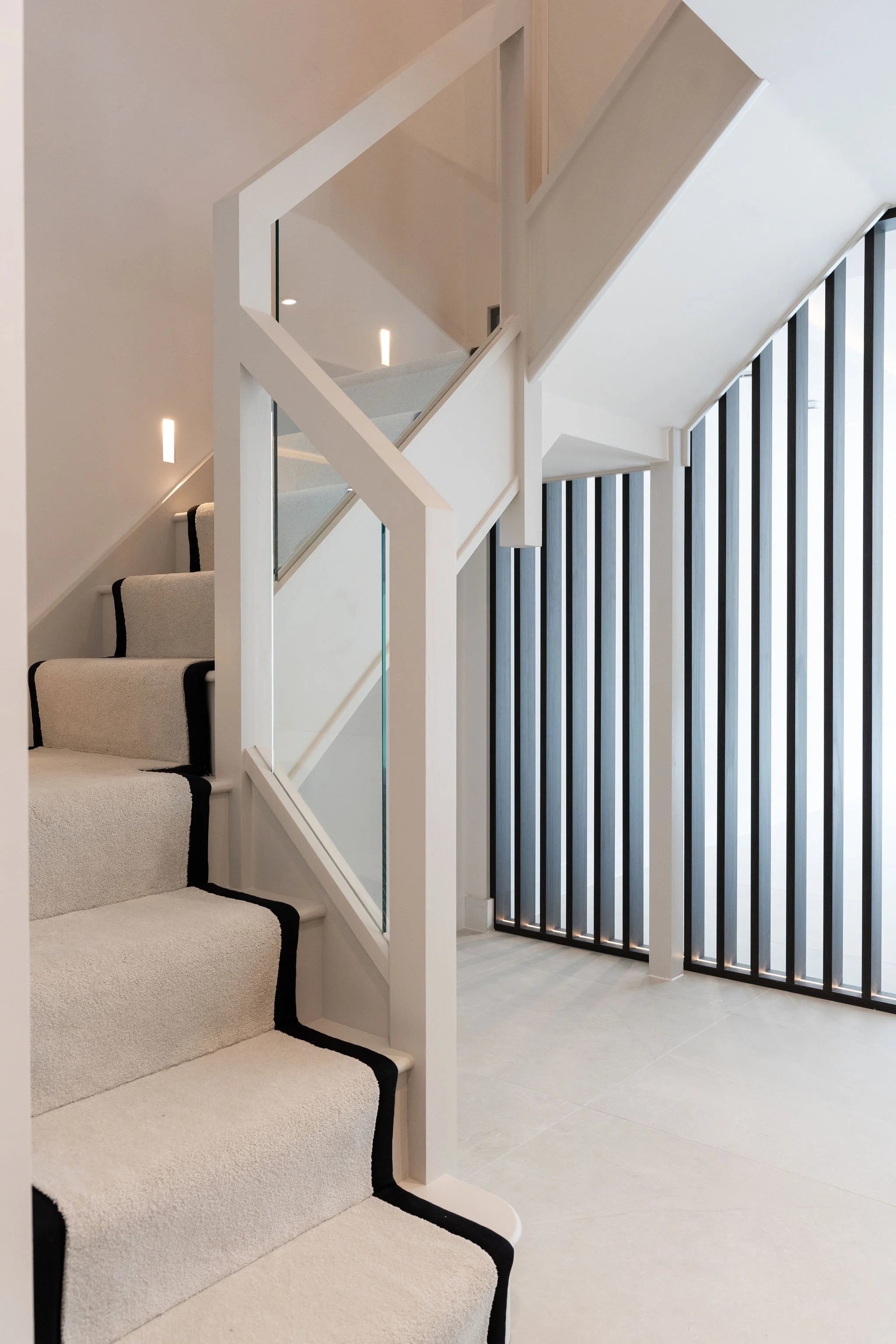Modern interior staircase with white walls, beige carpet with black trim, glass panel, and vertical black slat wall behind.