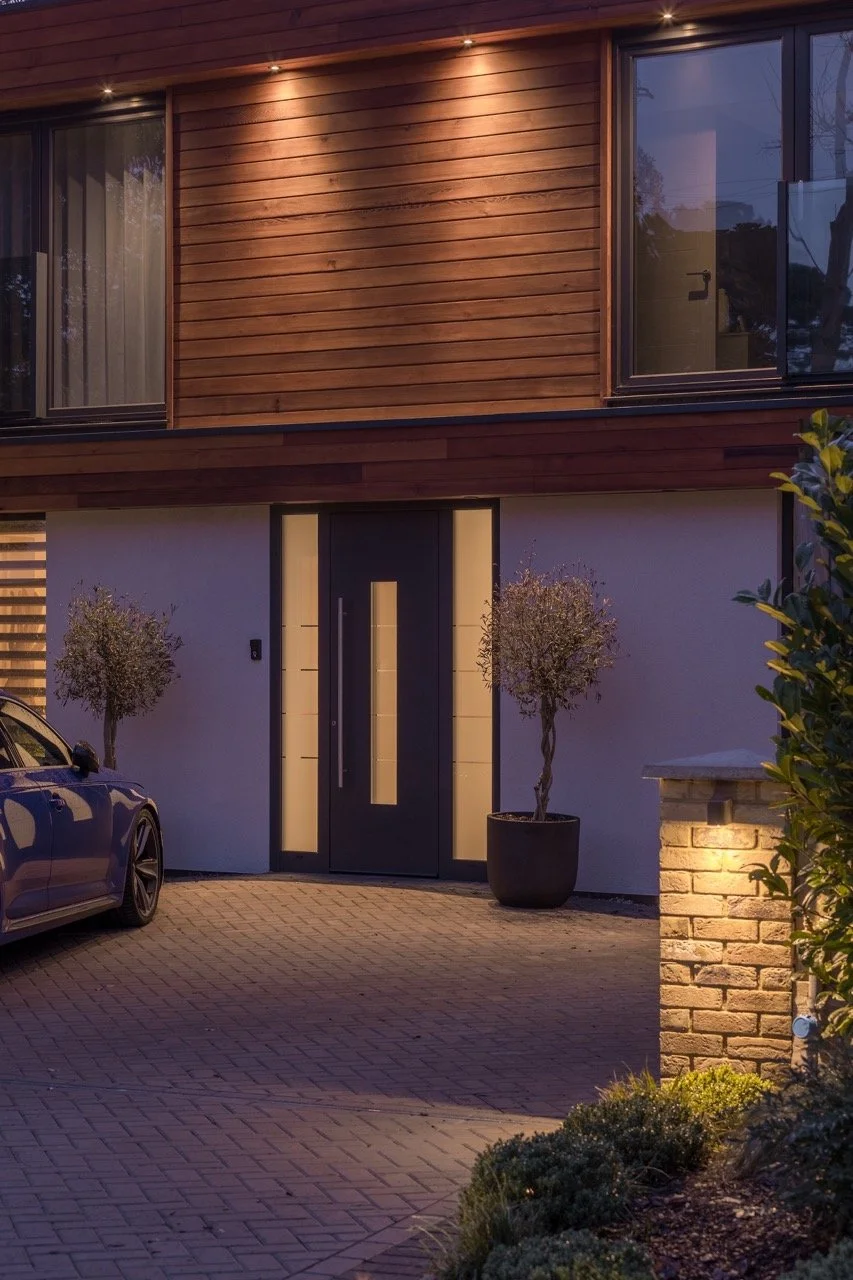 Modern house entrance with a black door, side windows, two potted trees, a brick mailbox, and a parked car, lit with outdoor lights in the evening.
