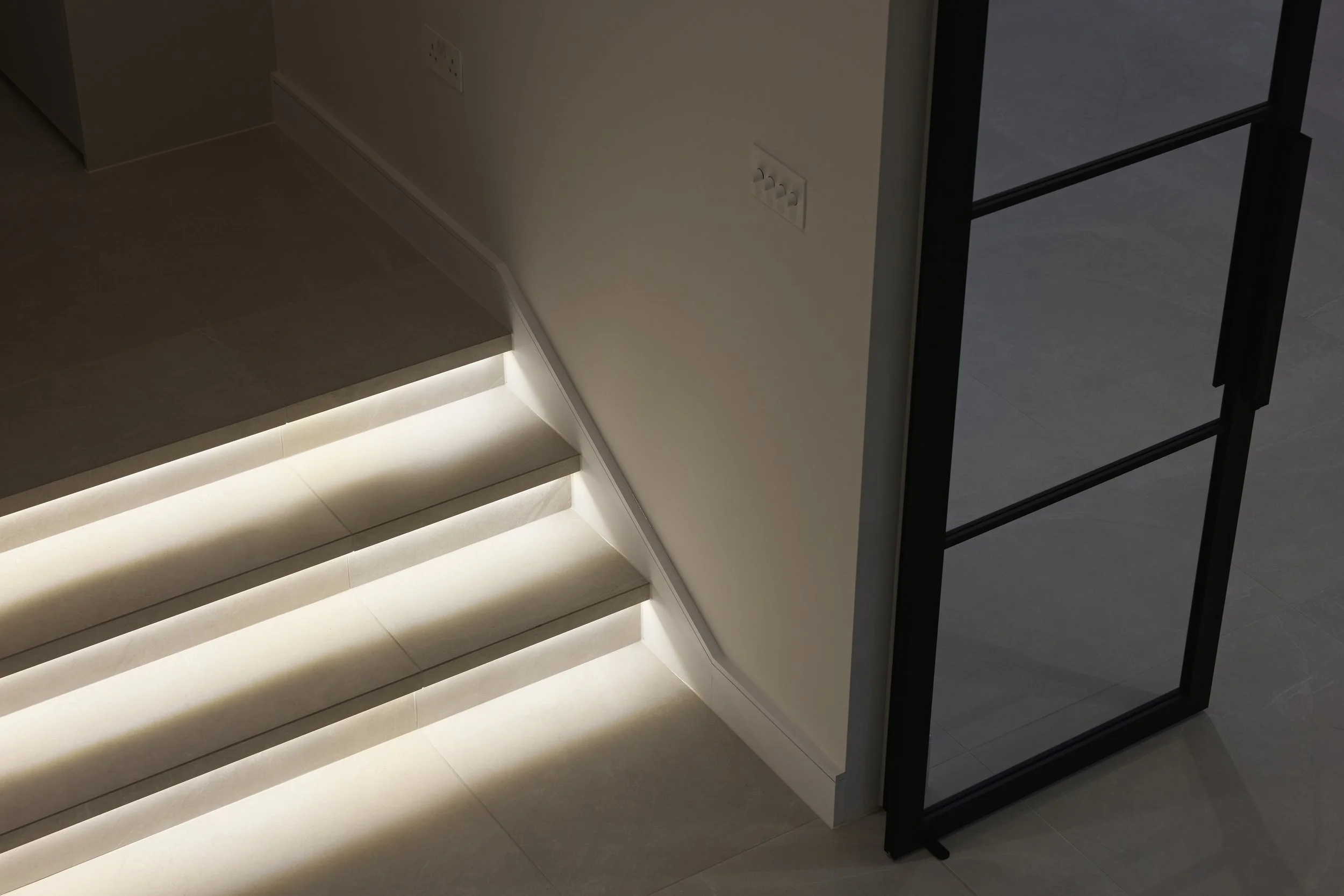 Modern interior staircase with illuminated steps, adjacent to a glass partition and a plain wall with electrical outlets.