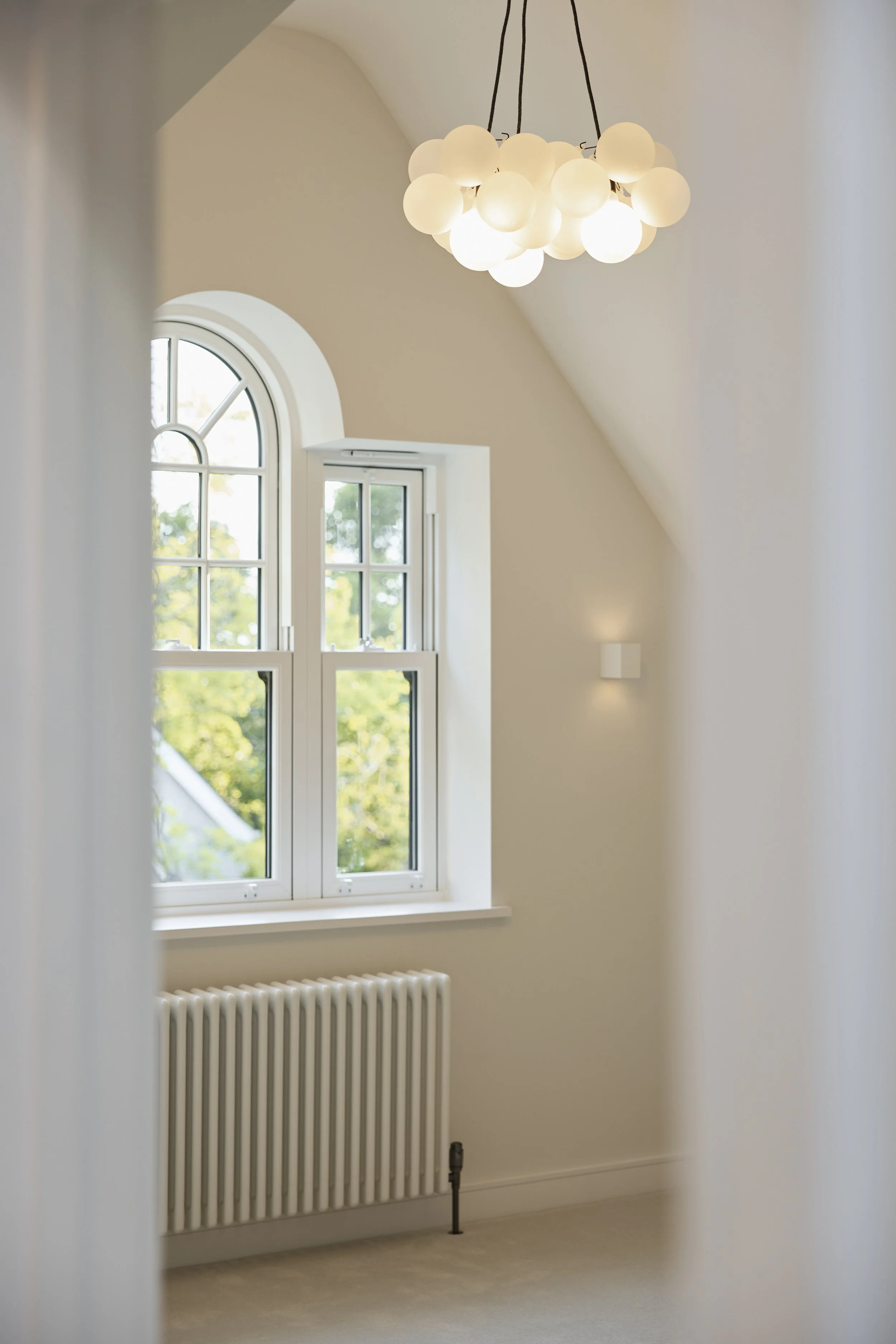 Bright room with arched window, modern chandelier with spherical bulbs, radiator, and wall-mounted light, viewed through a doorway.