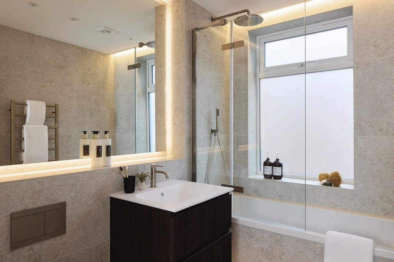 Modern bathroom with shower area, large mirror, frosted window, sink with black cabinet, and beige tiled walls.
