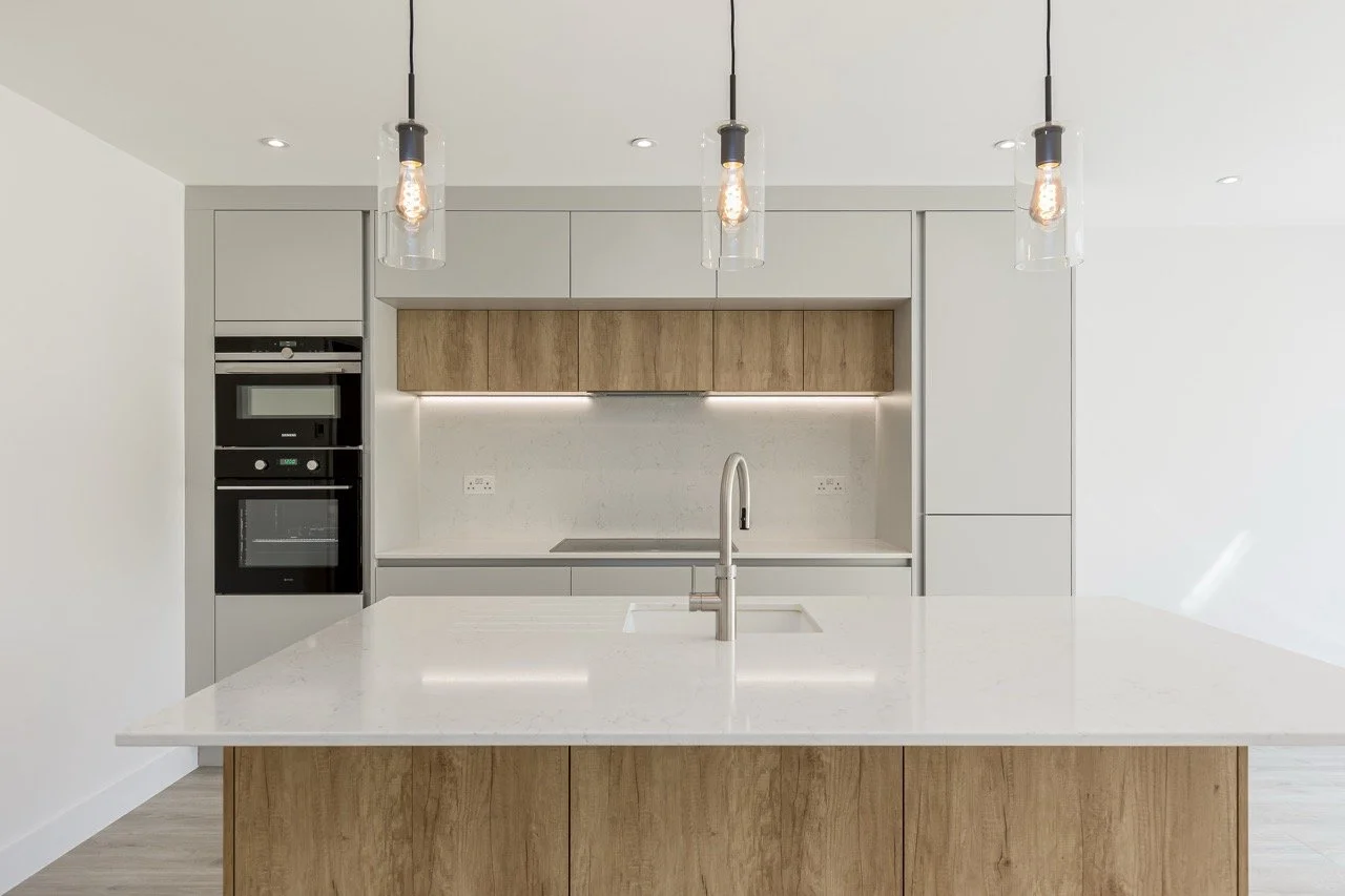 Modern kitchen with white countertops, wooden cabinets, built-in oven and microwave, and three pendant lights hanging above an island with a stainless steel faucet.