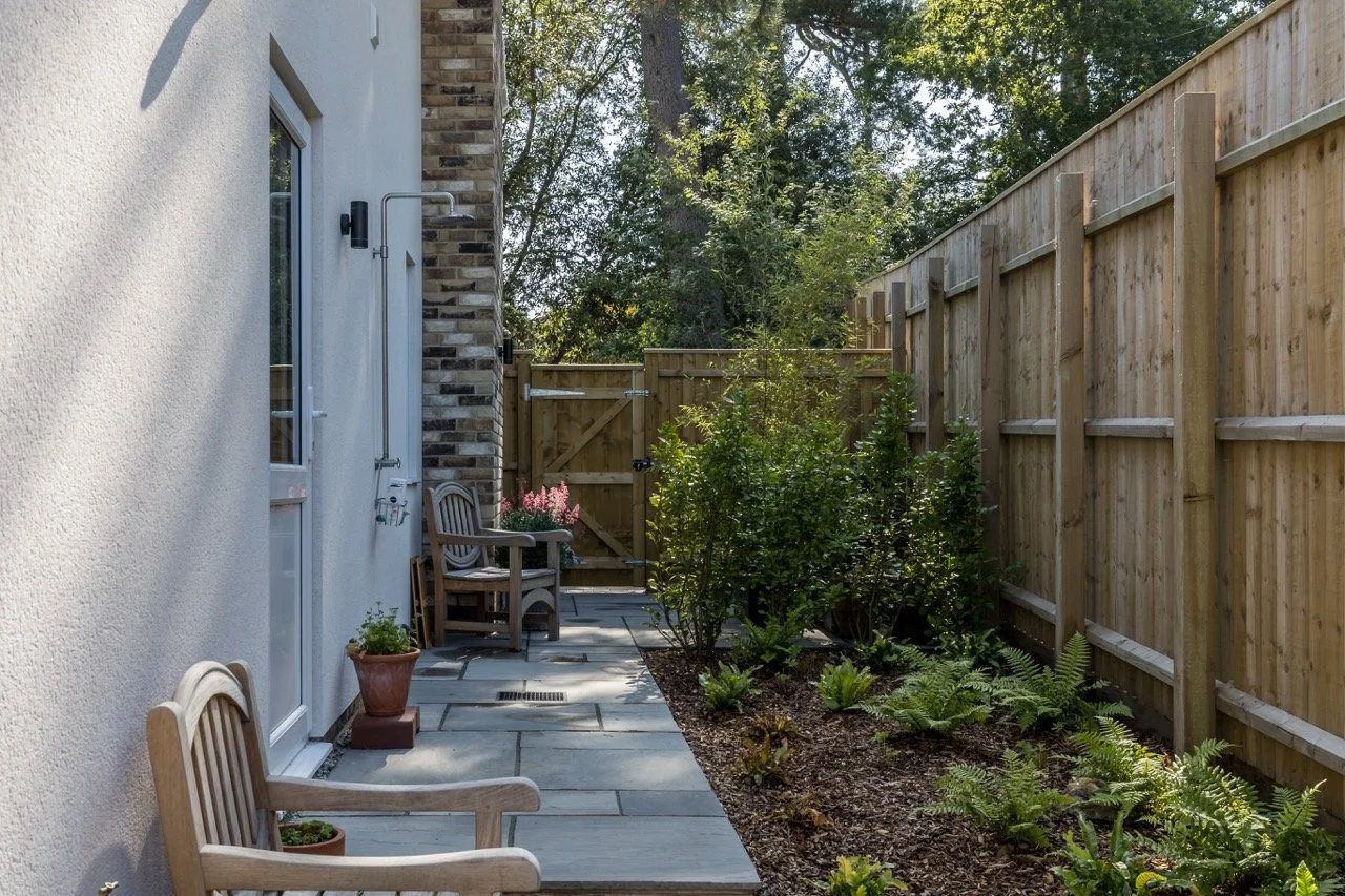 Small backyard patio area with two wooden chairs with potted plants on a stone-tiled pathway, next to a white house wall with outdoor shower, enclosed by a wooden fence, with trees in the background.
