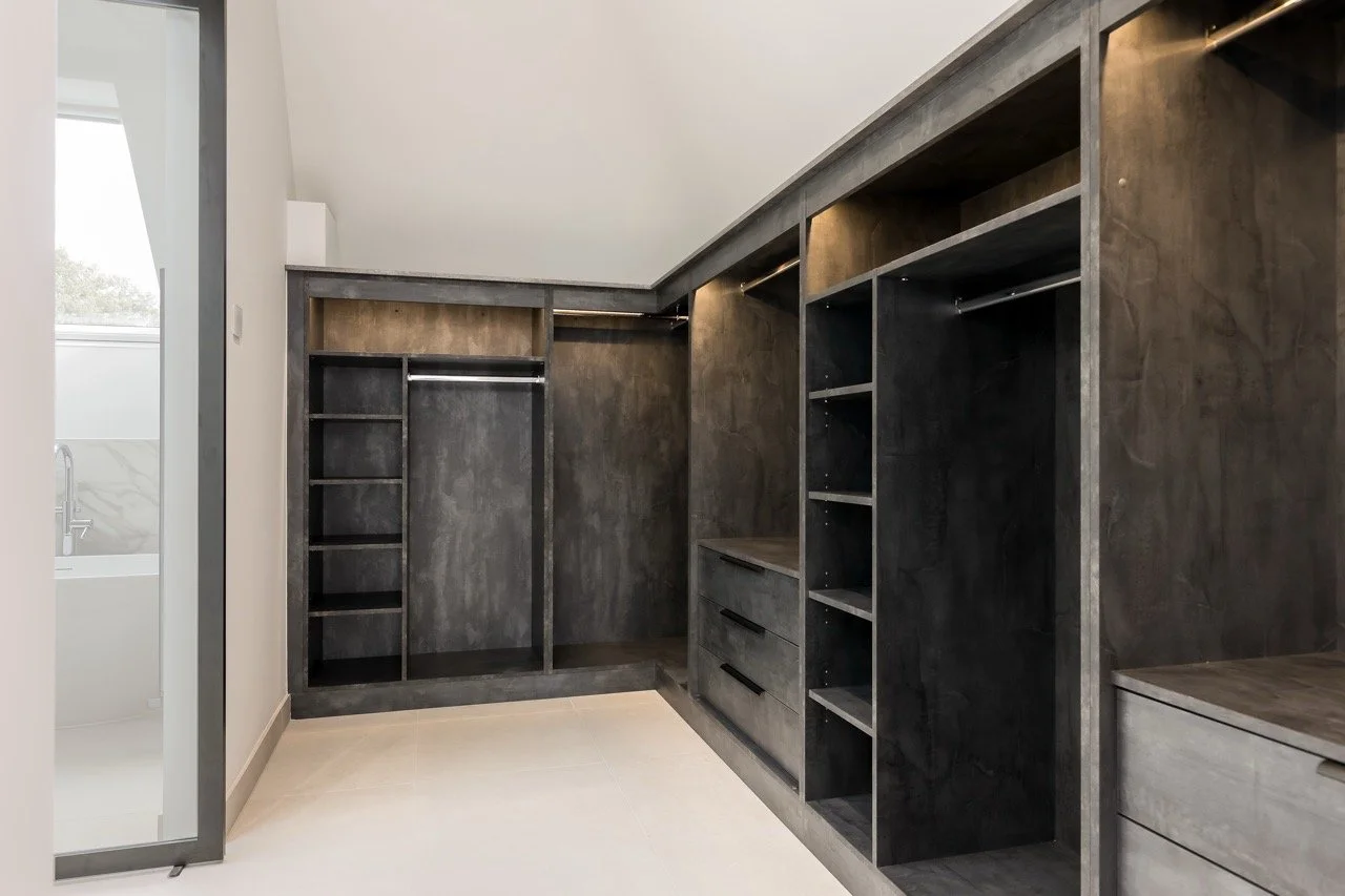 Empty walk-in closet with dark wood finish, open shelves, hanging rods, and drawers, adjacent to a bathroom with a bathtub.