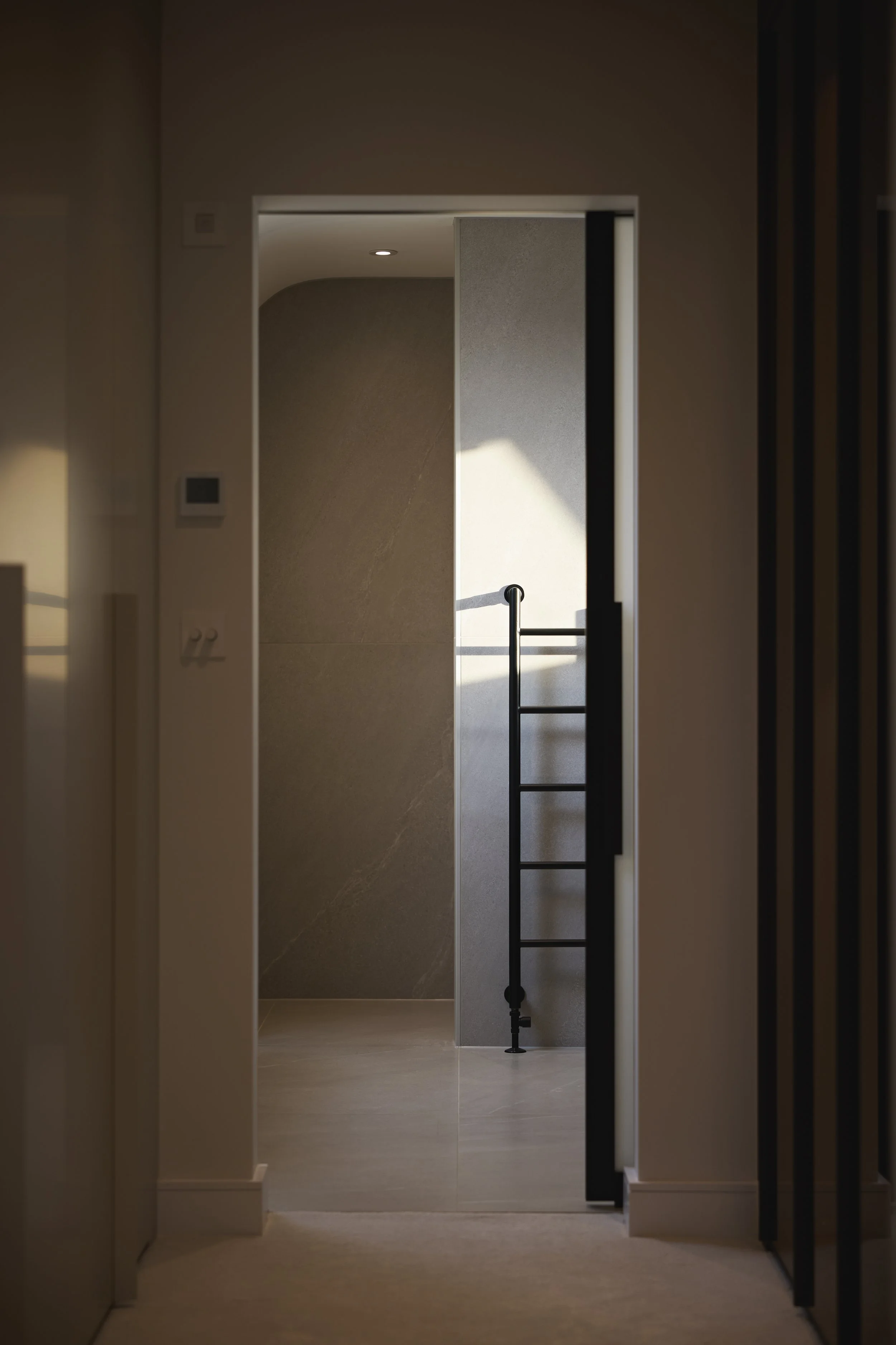 View through an open doorway into a room, with sunlight casting shadows on the wall, featuring a black ladder and a textured wall.