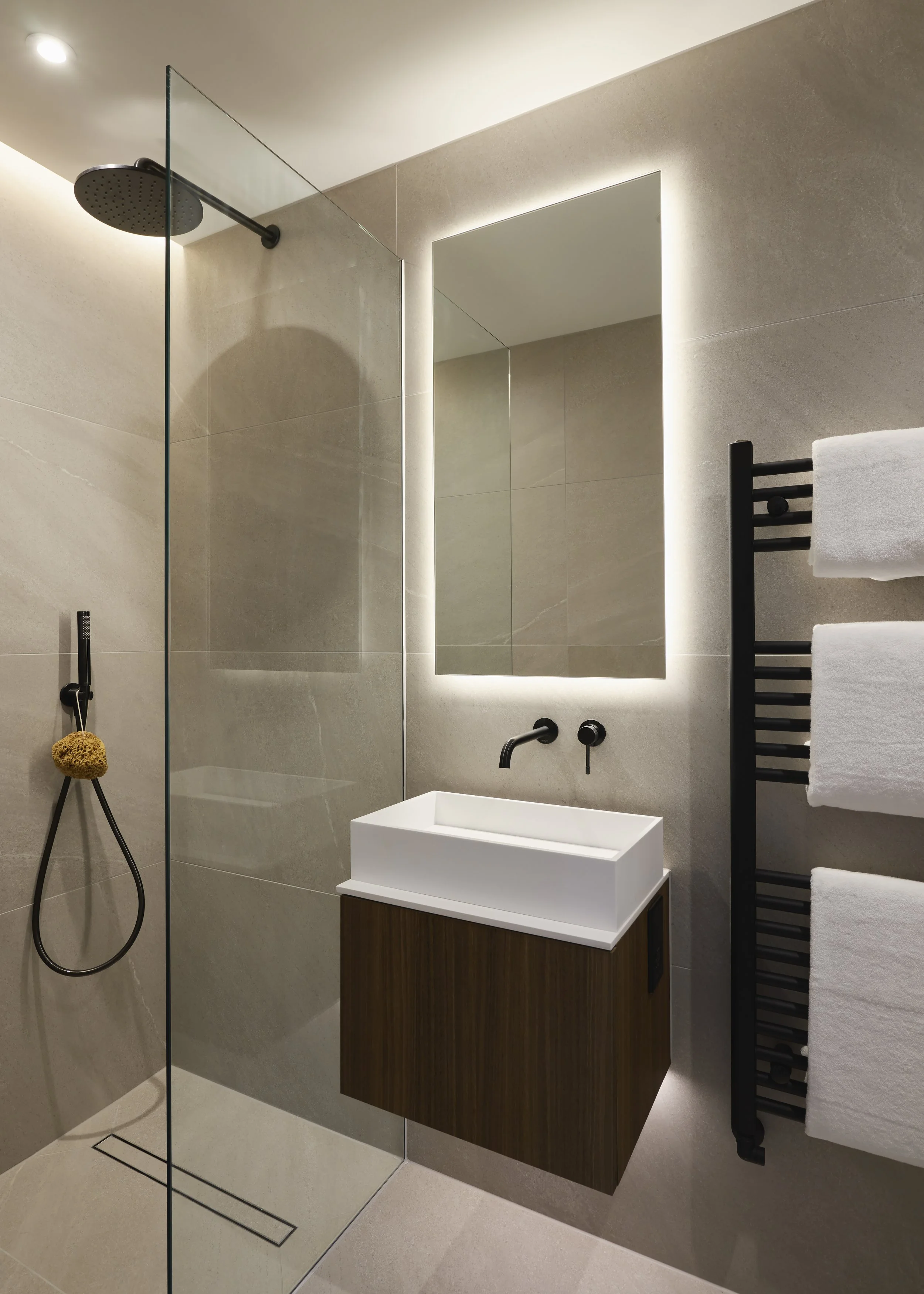 Modern bathroom with beige tiled walls, a glass shower enclosure, a rectangular white vessel sink on a wooden vanity, a large backlit mirror, and a black heated towel rail with white towels.