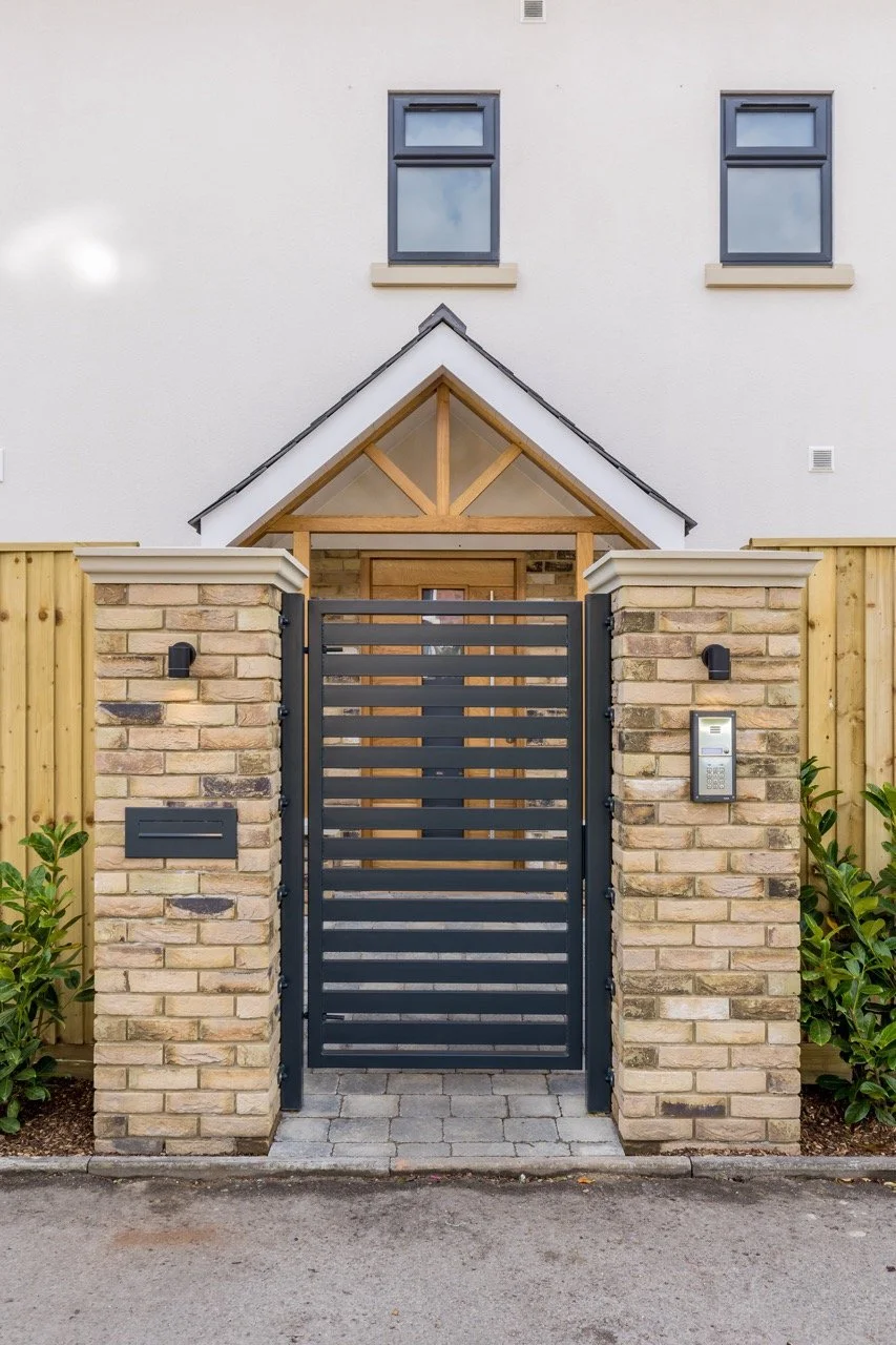 Modern house entrance with brick pillars, black metal gate, wooden features, and two upper windows.
