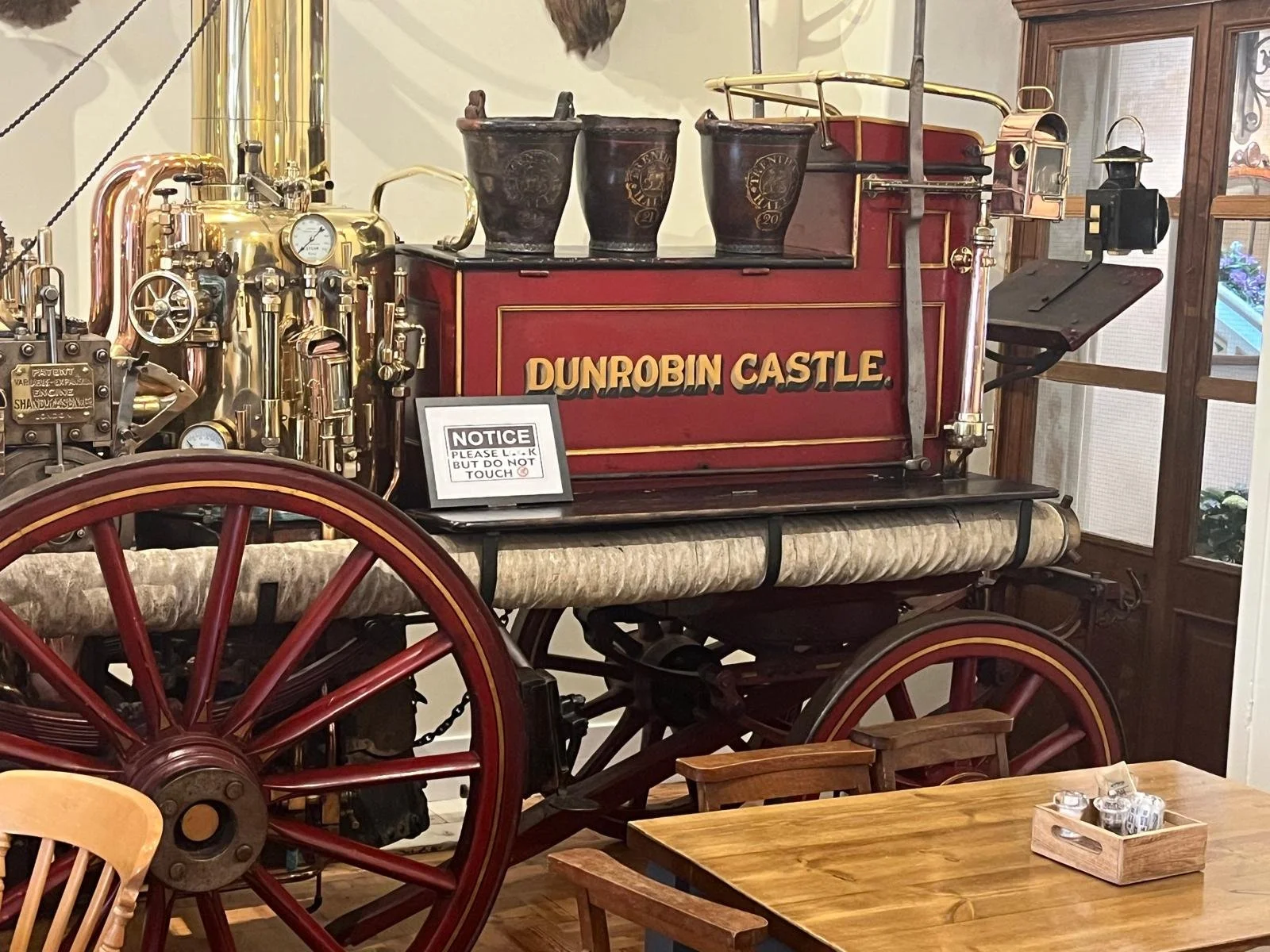 A vintage Dalnobrin Castle fire truck on display with a sign that reads 'NOTICE PLEASE LOOK BUT DO NOT TOUCH'. The fire truck features a red body with gold trim, three black pots on top, and a large brass engine on the side. The background includes a