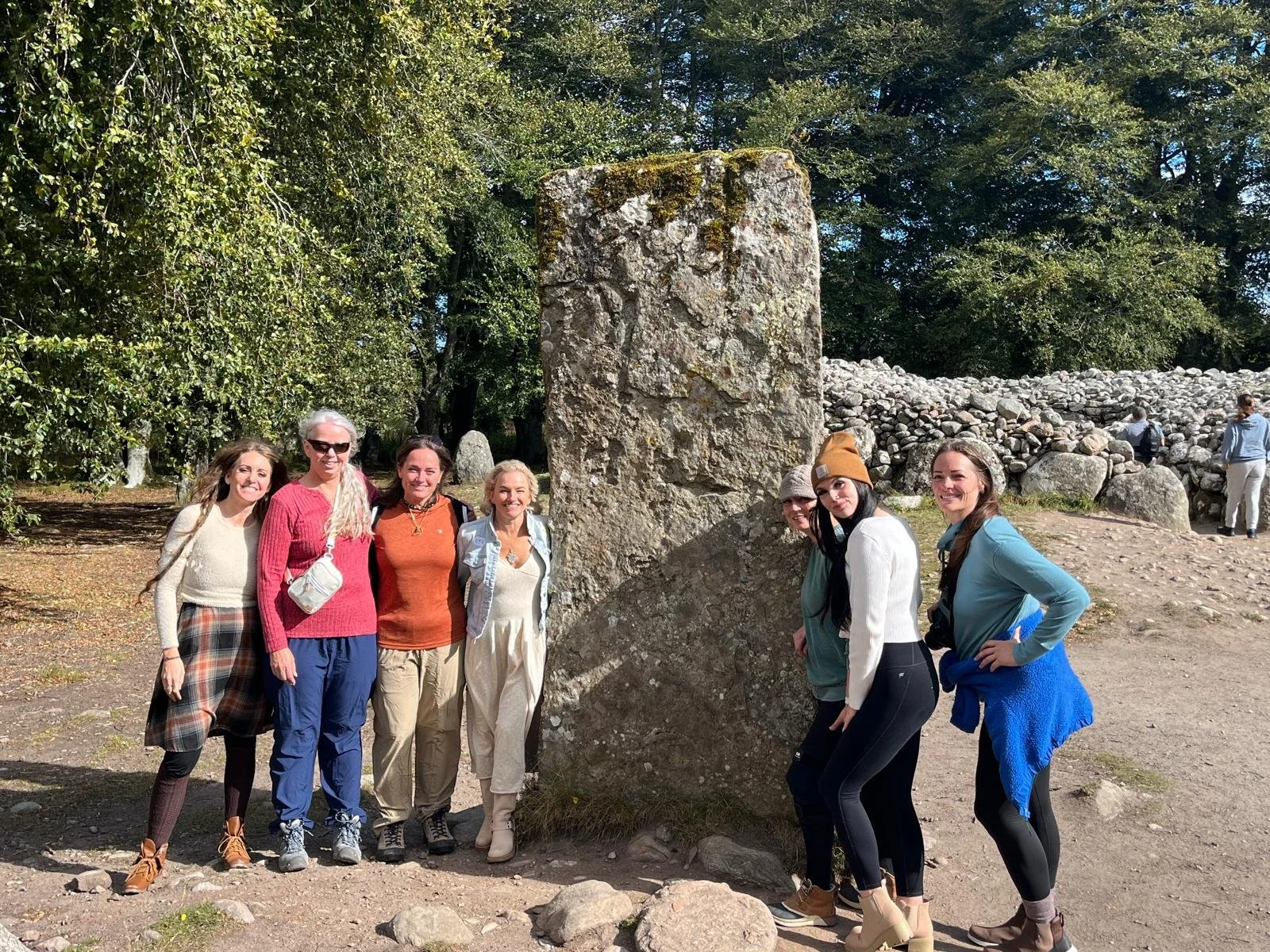Group of seven women and girls standing outdoors near ancient stone monument with a tall, rectangular stone and a small stone wall in the background, surrounded by trees.