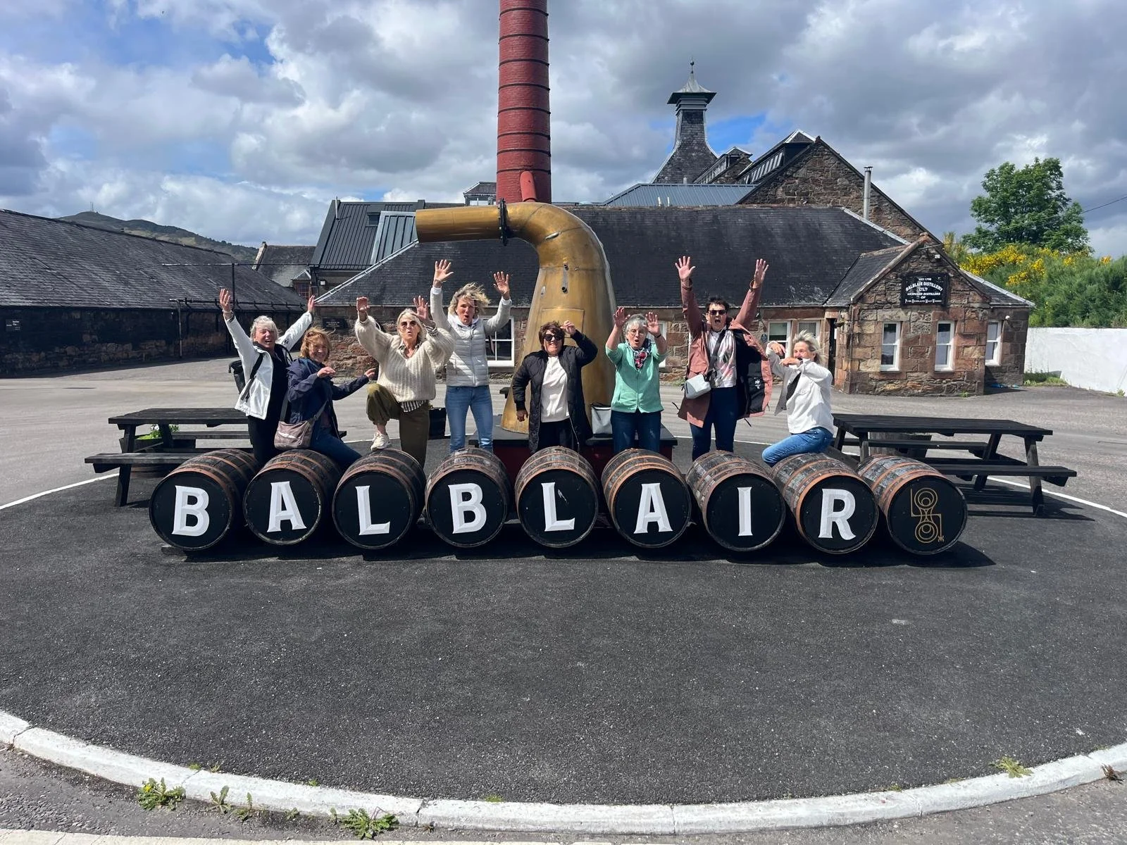 A group of people standing behind a row of barrels with letters spelling "BALBLAIR" in a town square, with a brown building and cloudy sky in the background.