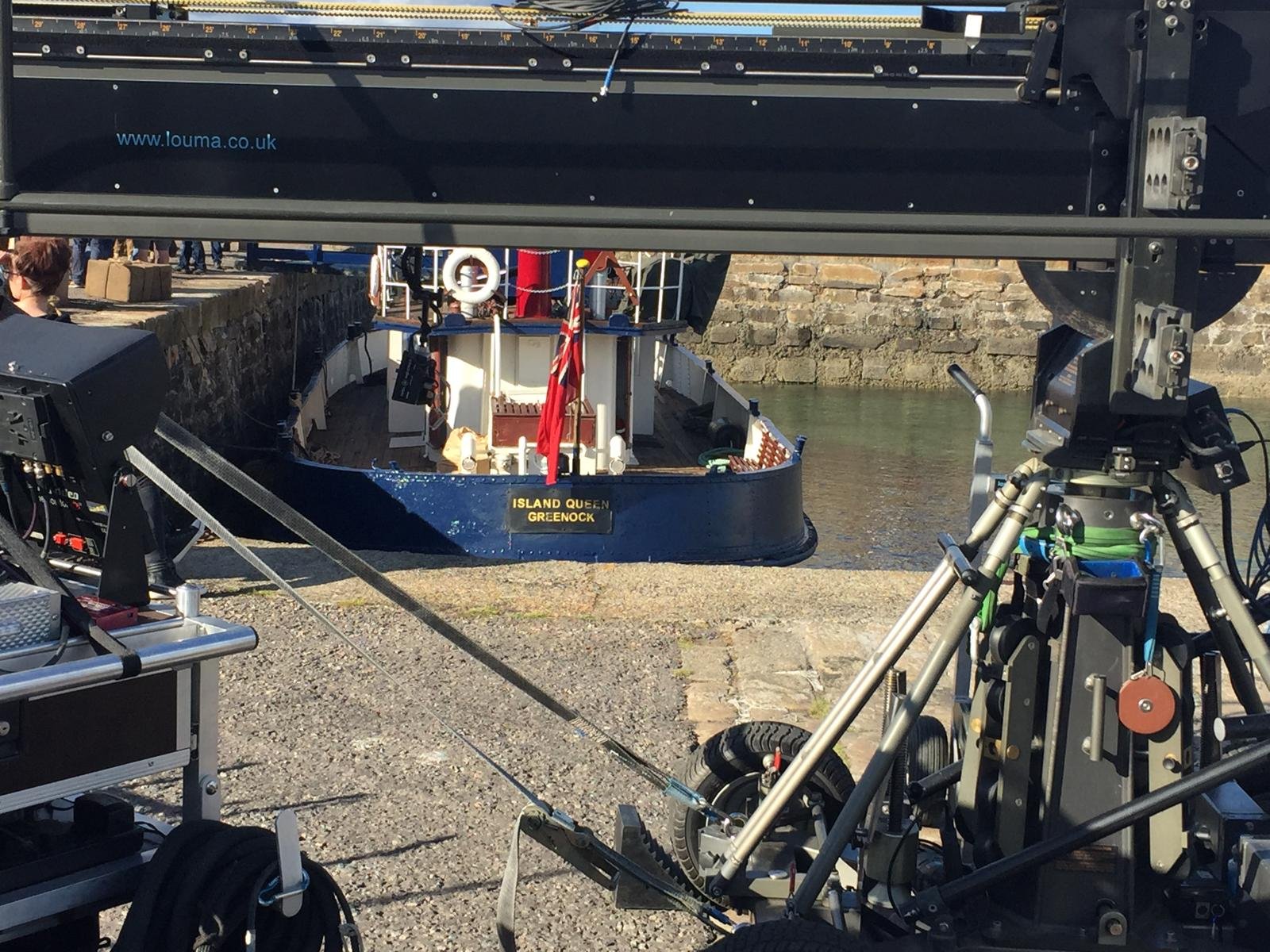 Camera equipment and a film set looking through a bridge or overhead structure at a small boat named 'Island Queen' in the water with brick walls and people on land in the background.