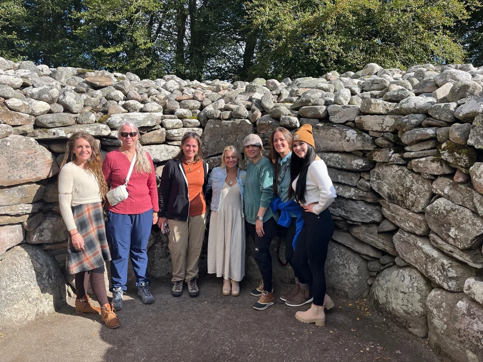 A group of seven women standing together outdoors in front of a large stone wall, smiling for the photo.