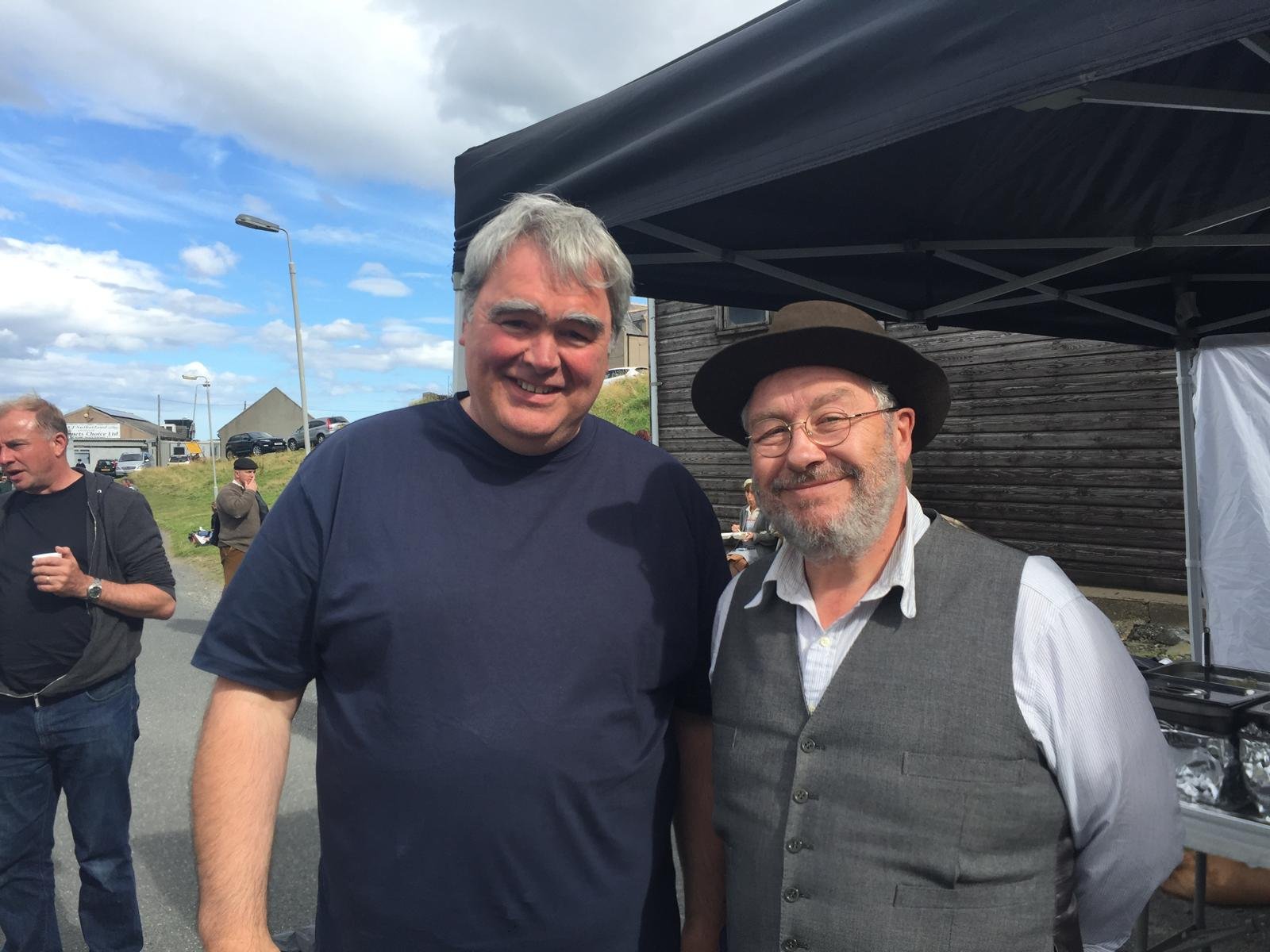 Two smiling men standing outdoors at a gathering, one wearing a dark t-shirt and the other in a light shirt, vest, and hat, with a tent and other people in the background.
