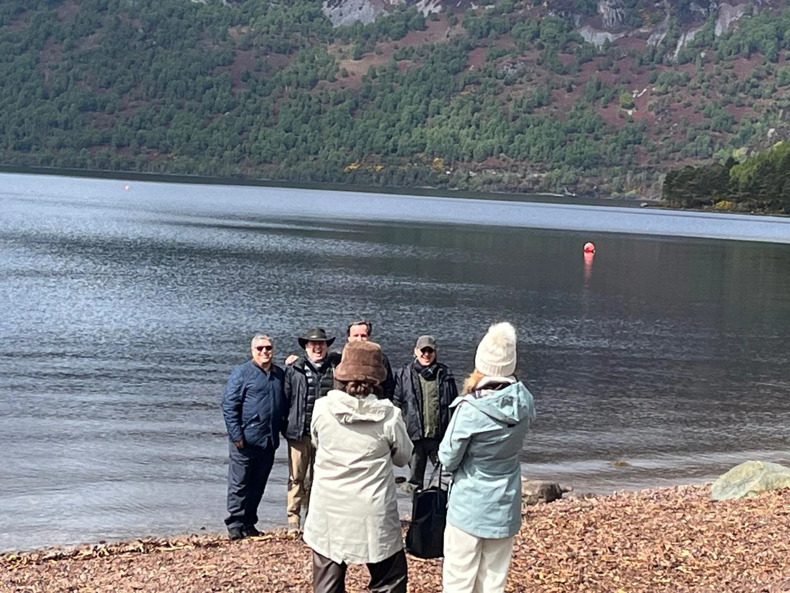 Four people are standing near a body of water, with hills and trees in the background. They are dressed in jackets and hats, and seem to be having a conversation.