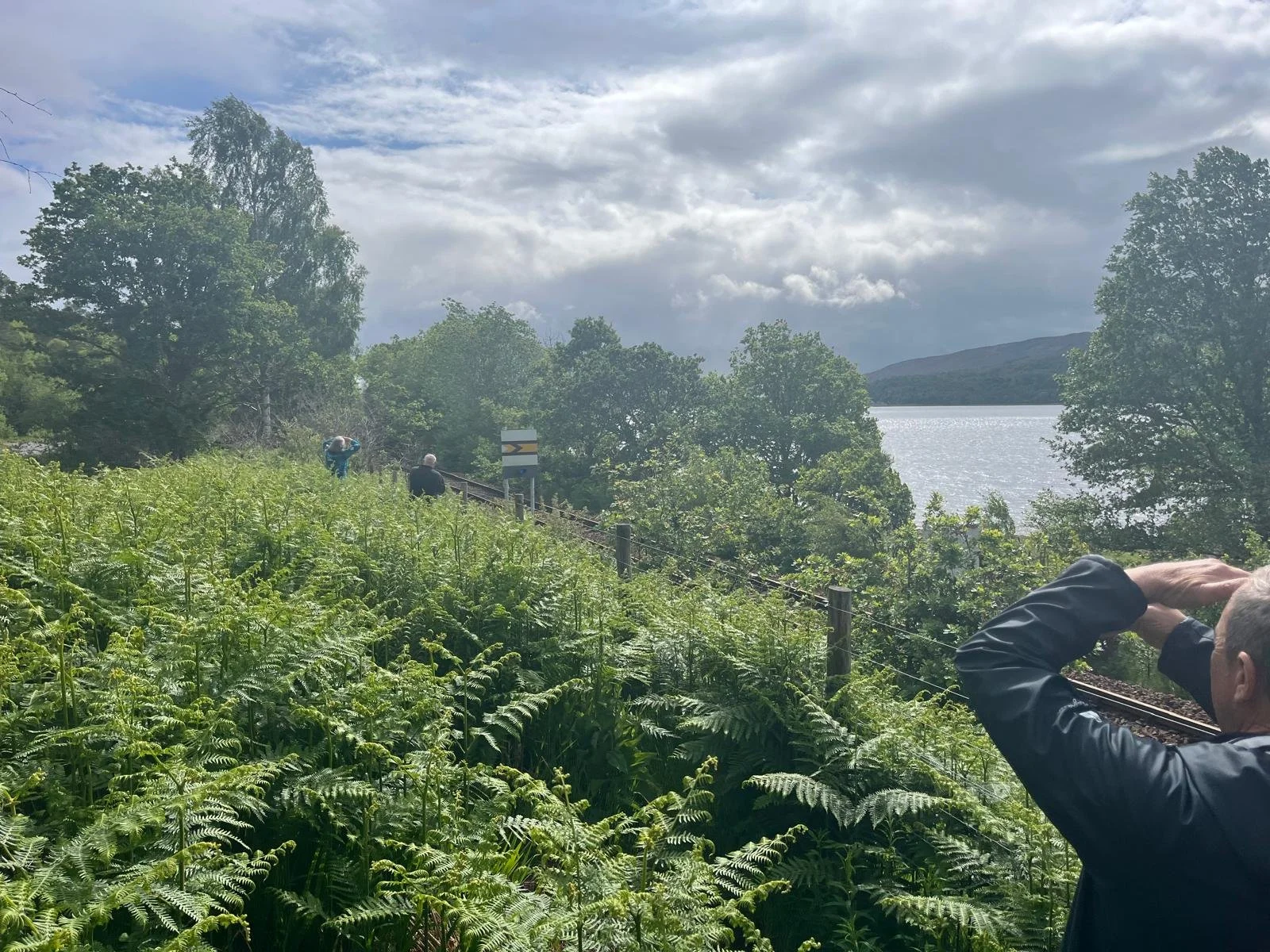 People standing on a railway track near a lake, surrounded by lush greenery and trees, with mountains in the background and cloudy sky.
